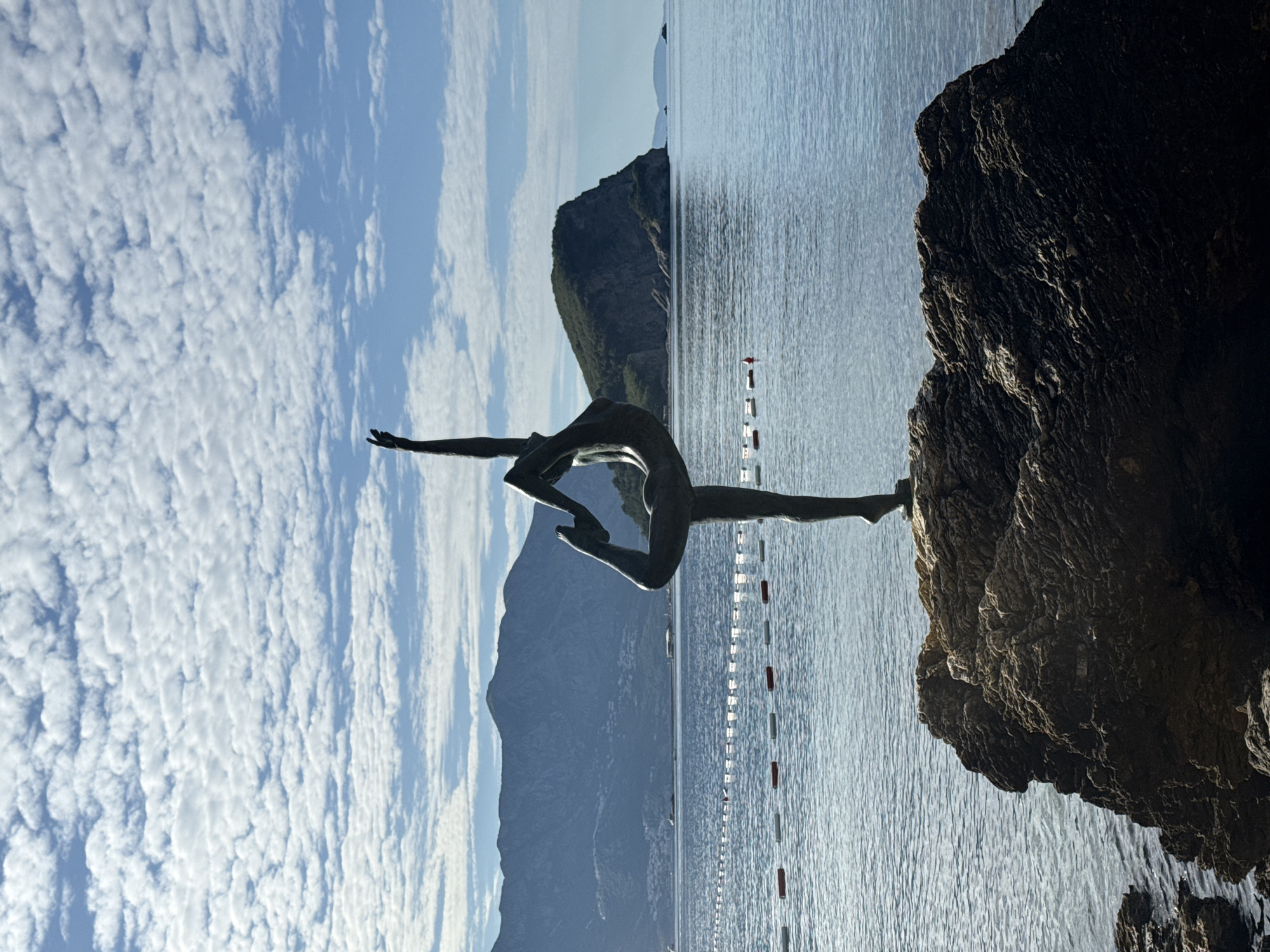 The image displays the "Dancer from Budva" statue, a prominent landmark in Budva, Montenegro. A bronze-colored statue of a female figure is centrally positioned, performing a balanced pose akin to a dancer's arabesque. She stands on one leg, with the other leg raised and bent backward, its foot held by the hand of the corresponding side, while her opposite arm extends vertically upwards. The statue is perched on a large, dark, rugged rock formation emerging from the calm, blue-grey Adriatic Sea in the foreground. In the mid-ground, a line of white and red buoys stretches across the water. The background features a distant range of dark, mountainous terrain with visible vegetation and scattered structures to the left, and a prominent, greener, rocky island or headland to the right. The sky above is light blue and populated with numerous small, fluffy white clouds.