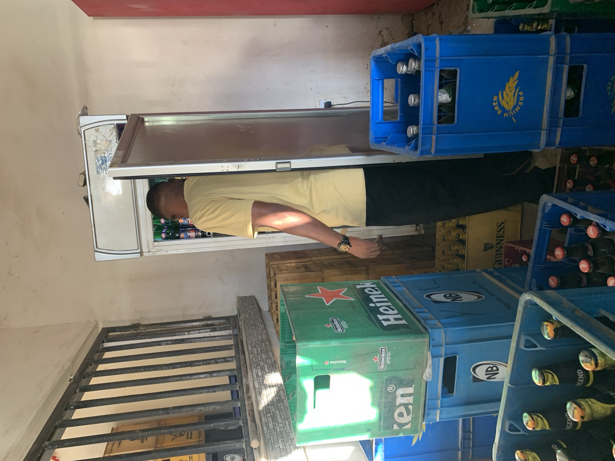 This image captures an indoor stockroom or retail storage area in New Stadium, Nigeria.
A male individual, seen from behind and partially obscured by an open refrigerator door, stands centrally. He wears a short-sleeved yellow t-shirt and dark trousers. A gold-toned watch is visible on his left wrist. He is actively engaged with the open, silver-colored commercial display refrigerator, which contains multiple bottled beverages. The top section of the refrigerator shows minor damage or repair with clear adhesive. An electrical cord extends from the refrigerator to a white wall outlet on the right wall.
The environment is characterized by stacked beverage crates. In the immediate foreground, a blue plastic crate holds bottles with gold-foil caps and "LEGEND" branding. Directly behind it, a prominent green plastic crate with "Heineken" branding and a red star logo is stacked on two blue plastic crates marked "NB" (Nigerian Breweries Plc) with an oval logo. Further right, stacked blue crates feature a yellow "PREMIUM BREW" and wheat stalk emblem, with bottles displaying silver caps. Another blue crate on the far right holds bottles with red caps. In the mid-ground, behind the green Heineken crate, a stack of brown or yellowish crates, one clearly labeled "GUINNESS," is visible alongside wooden pallets.
To the left, a window opening is secured by vertical metal bars, below which a tiled ledge is present. Beyond the bars, additional stacked crates are faintly visible. The walls are painted a light color, exhibiting signs of wear, dirt, and minor discoloration, particularly near the uneven, dirty concrete floor. A section of wall on the far right is painted red. The scene suggests a functional, possibly high-traffic, storage area for beverages.