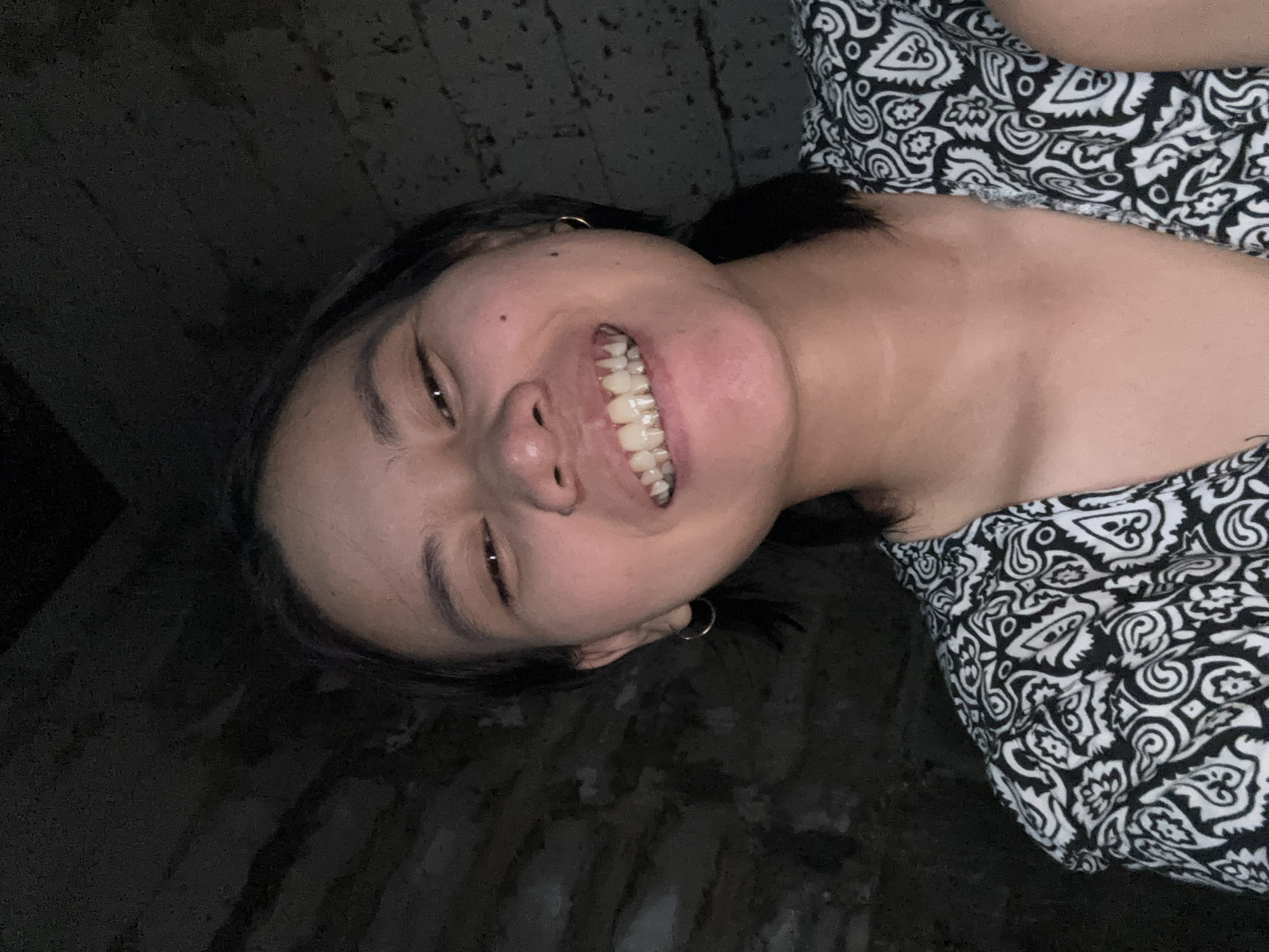 A young woman with dark hair and fair skin smiles at the camera. She is wearing a black and white patterned top and hoop earrings. The background is dark and appears to be a textured wall. The image was taken in East Jakarta, Indonesia.