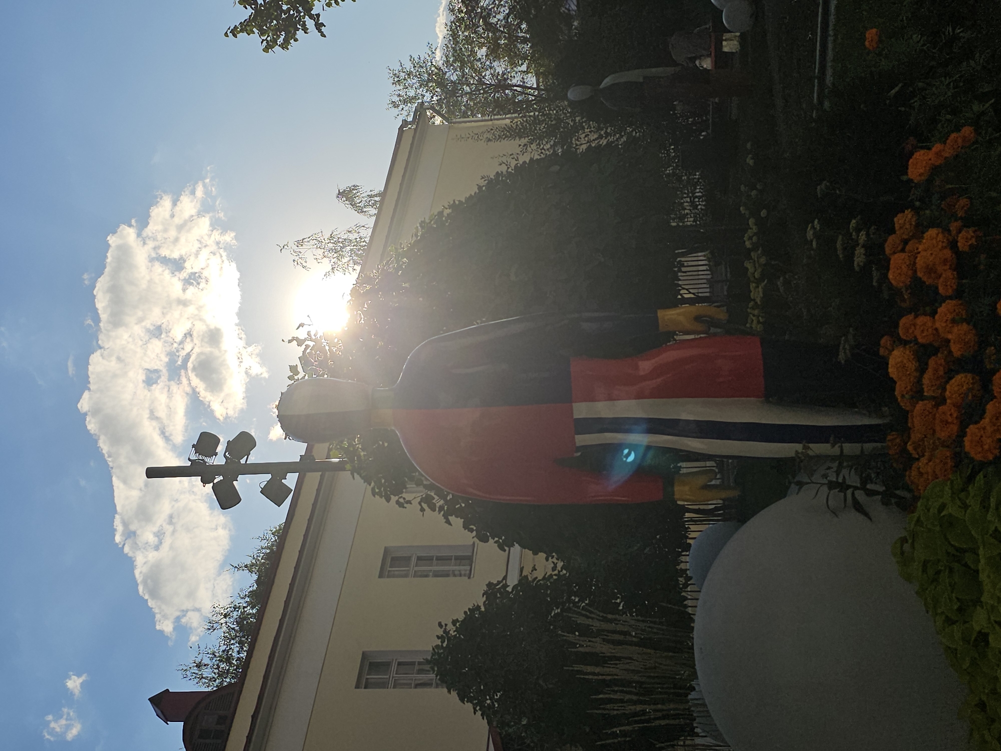 This is a medium-angle, outdoor photograph taken in Moscow, Russia, likely in an urban park or garden. The scene is illuminated by bright sunlight, with the sun partially obscured by a large cloud.
In the foreground, a large, colorful statue of a person is prominent. The statue is divided vertically into red and black halves on its torso. The lower body of the statue features vertical stripes of red, white, and blue, with yellow accents on the hands. The statue is positioned in a garden setting, with vibrant orange flowers in the lower right corner and lush green foliage and tall grass-like plants behind it. A large, smooth, grey, spherical object is partially visible in the lower left quadrant of the image.
In the mid-ground, a building with a pale yellow facade and several windows is visible behind the statue. Trees with dark green foliage partially obscure the building and the statue. To the left of the statue, a tall, dark pole with multiple spotlights or lamps is erected.
In the background, additional trees and foliage are present. To the right of the main statue, a fainter, smaller, light-colored statue of a person is visible, also appearing to be in a garden. The sky above is a clear blue with a few white clouds, and the sun is a bright glare visible through the cloud and trees. The overall impression is of a public outdoor space with artistic installations and natural elements, bathed in warm sunlight.