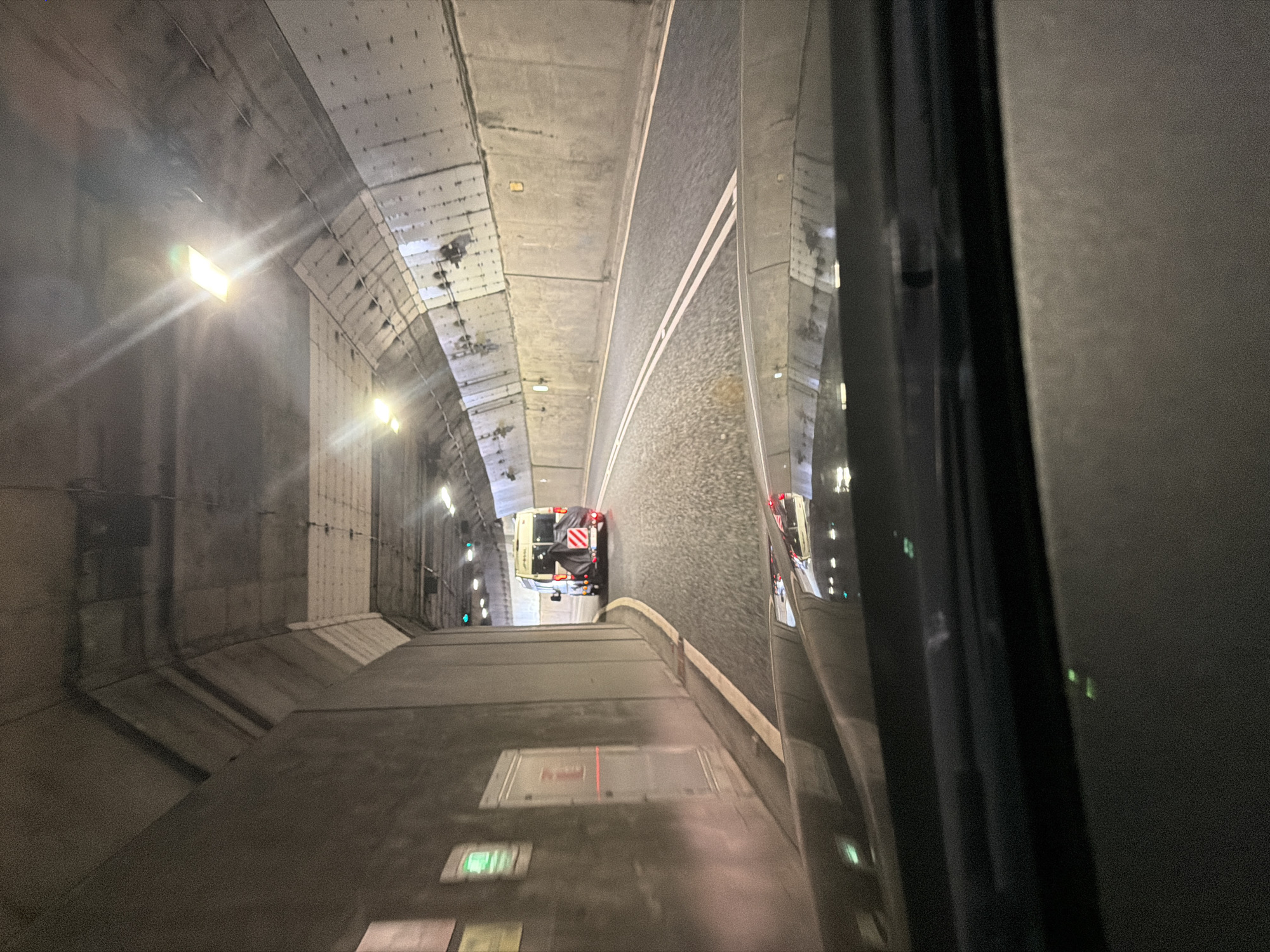The image captures an interior view of a concrete tunnel, taken from within a vehicle. The perspective looks forward and slightly downward, with a dark, reflective surface on the right side of the frame, possibly a window, which mirrors the scene ahead.
The tunnel's structure consists of concrete walls and ceiling, featuring visible panels and fixtures. Bright artificial lights illuminate the space, with some lights producing noticeable lens flares. Along the left wall, several green lights are visible, possibly signaling or safety indicators.
A roadway or track occupies the center of the tunnel, characterized by a dark, textured surface and distinct double white solid lines that separate two lanes. Ahead, a light-colored vehicle, resembling a maintenance van or small truck, is positioned on the track. Its rear is equipped with prominent red and white diagonal striped reflective markings, and a black cover or equipment is visible.
The foreground on the left shows part of the viewing vehicle's interior or floor, with blurry rectangular lights, one appearing reddish and another greenish. No readable text is discernible anywhere in the image. The scene suggests travel through an enclosed environment, likely a tunnel at night or indoors, within the North Tyneside area of the United Kingdom, possibly a part of a railway or road system.