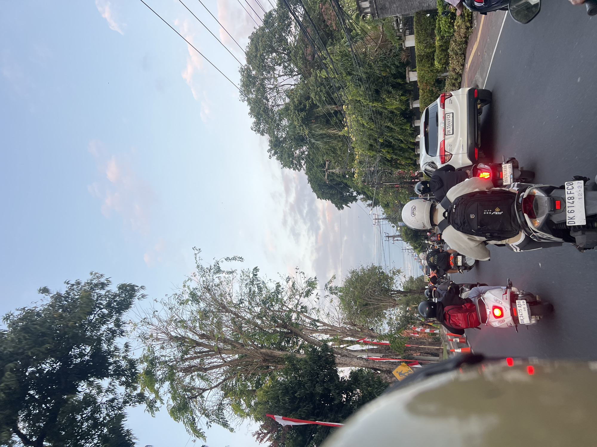 The image depicts a traffic scene in Denpasar, Indonesia, likely during the late afternoon or early evening, given the soft light and scattered clouds in the sky. The foreground is dominated by the backs of several motorcycles, with riders wearing helmets and backpacks. One white scooter in the middle ground has a visible license plate, "DK 3475 ADX". Behind the motorcycles, a white SUV is visible in the right lane, also with a license plate, "DK 1564 FCK".
The road is paved, and traffic appears to be moving slowly. On the left side of the road, there are trees lining the street, with some adorned with Indonesian flags. Orange traffic cones are also present along the roadside. The background features dense foliage, power lines, and the sky. The overall impression is one of urban congestion.
