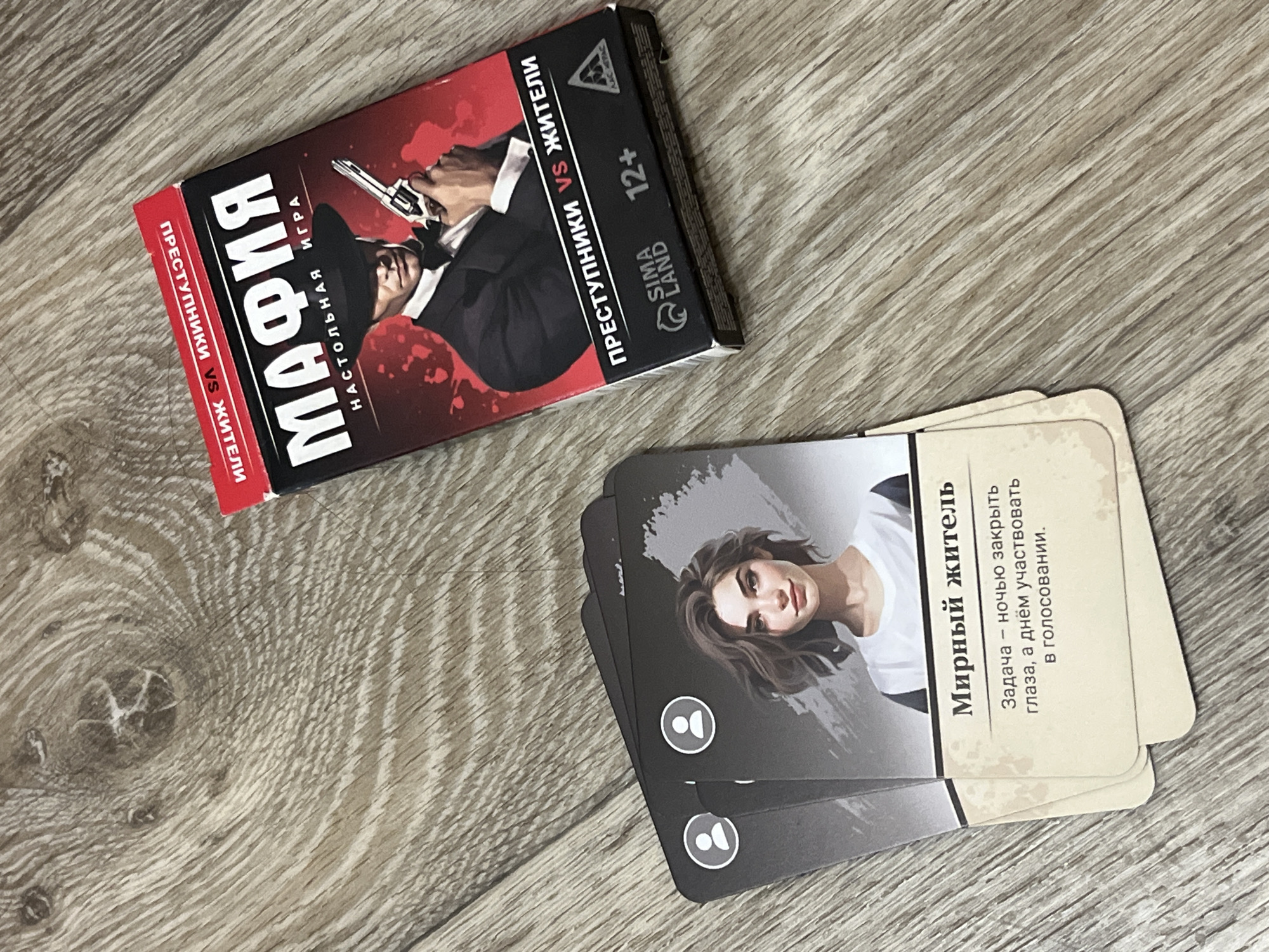 The image displays a card game titled "Mafia" and several character cards on a wooden surface. The "Mafia" game box is predominantly red and black, featuring an illustration of a man in a fedora holding a handgun. Text on the box includes "МАФИЯ" and "НАСТОЛЬНАЯ ИГРА" (Mafia, Board Game), along with "ПРЕСТУПНИКИ VS ЖИТЕЛИ" (Criminals vs. Residents) and "12+". The character cards are black and white, with one card prominently showing a portrait of a young woman and the title "Мирный житель" (Peaceful Resident). Below the title, there is a description of the character's role: "Задача – ночью закрыть глаза, а днём участвовать в голосовании." (Task - close your eyes at night, and participate in voting during the day.). The wooden surface appears to be a floor with a wood grain pattern. The location of Дачное, Russia, is not depicted or indicated in the image.