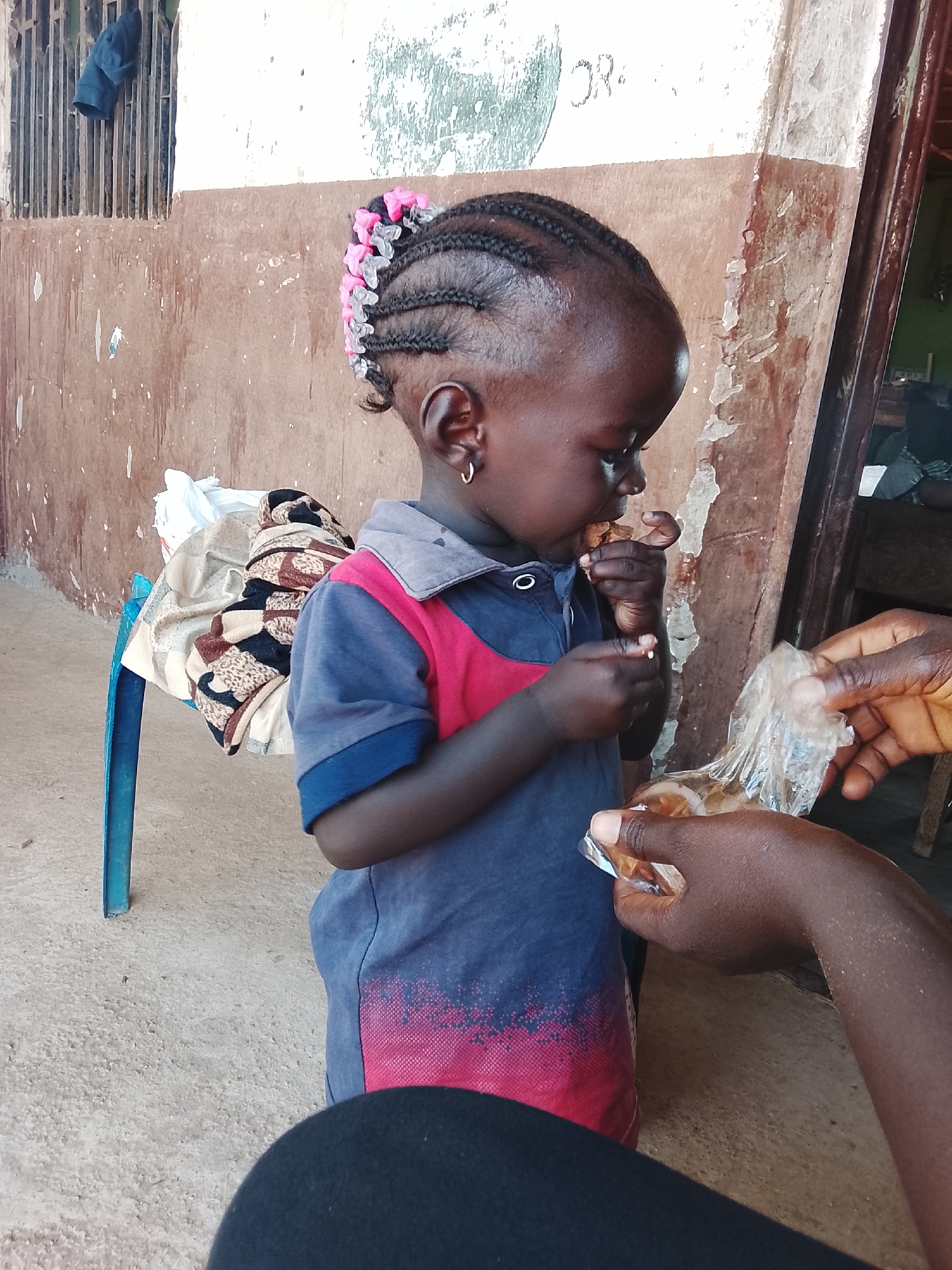 A young child, likely a toddler, is being fed by an adult's hands from a clear plastic bag. The child has braided hair adorned with pink and white beads and is wearing a two-toned polo shirt, blue and red, with a red, pixelated design on the lower front. The child is looking down and eating something from their own hands. The scene appears to be outdoors, on a concrete surface. Behind the child, a blue plastic chair is visible, covered with patterned fabric and a white plastic bag. The background consists of a weathered, brown and cream-colored wall with peeling paint and some faint markings or writing. To the left, a metal grate with a dark blue garment hanging on it is present. To the right, the wall has a dark wooden frame. The overall impression is a candid moment of a child being given a snack in an informal, possibly residential, setting. The location is identified as the city of Akwanga, Nigeria.