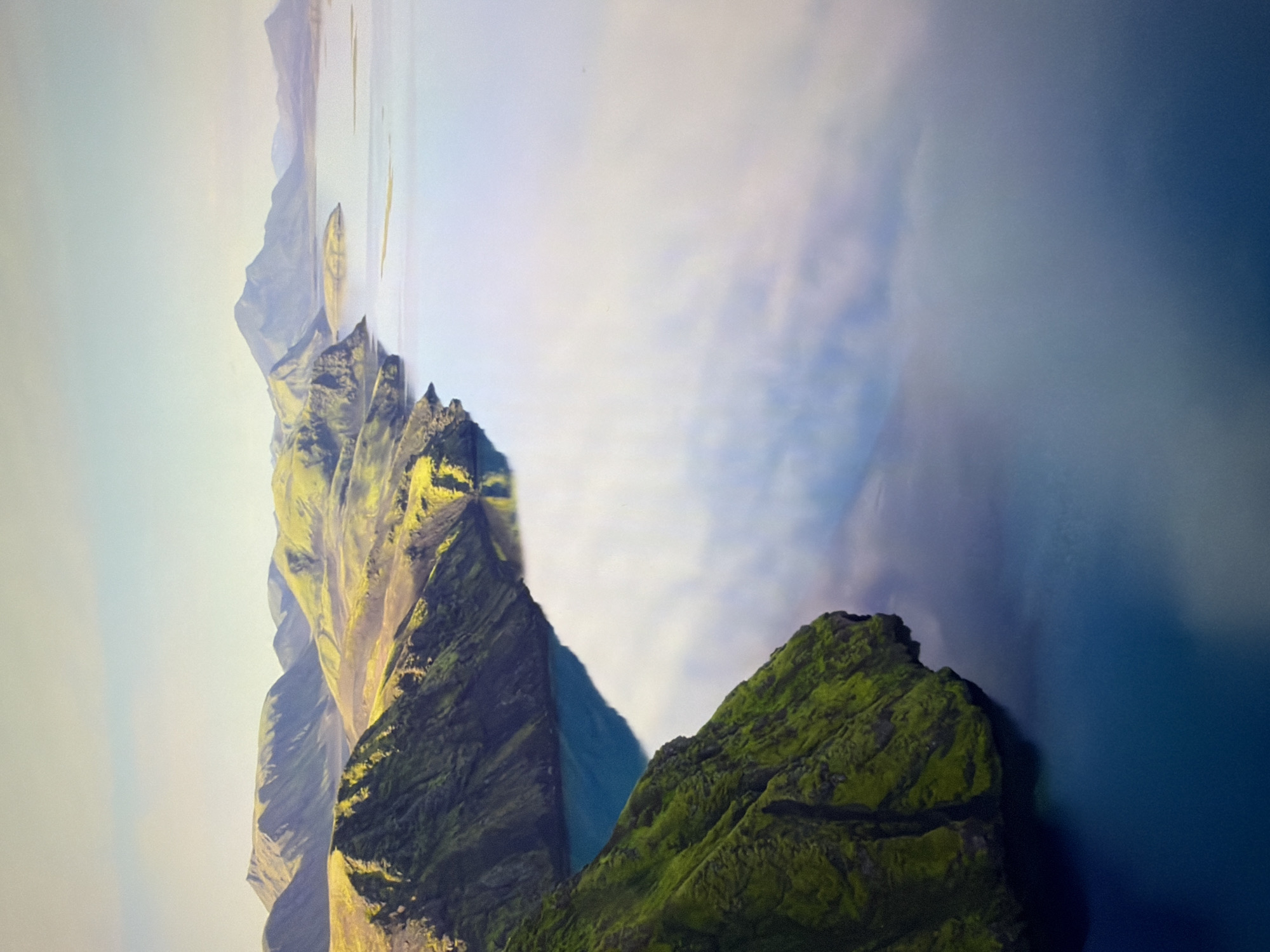 The media file displays a dramatic landscape with a prominent, rugged mountain range covered in lush green moss and vegetation. The mountains are sharply defined with steep slopes and craggy peaks, catching the sunlight in places, creating a contrast between light and shadow. To the right of the mountain range, a vast expanse of deep blue water stretches out, reflecting the soft, hazy sky. Wisps of white clouds or fog are present in the sky and partially obscure the distant mountains, adding a sense of depth and mystery to the scene. The overall impression is one of a wild, untamed natural environment. The perspective suggests an aerial view, looking down and across the landscape. The lighting indicates it could be either early morning or late afternoon, with the sun casting long shadows. There is no visible text in the media file.
