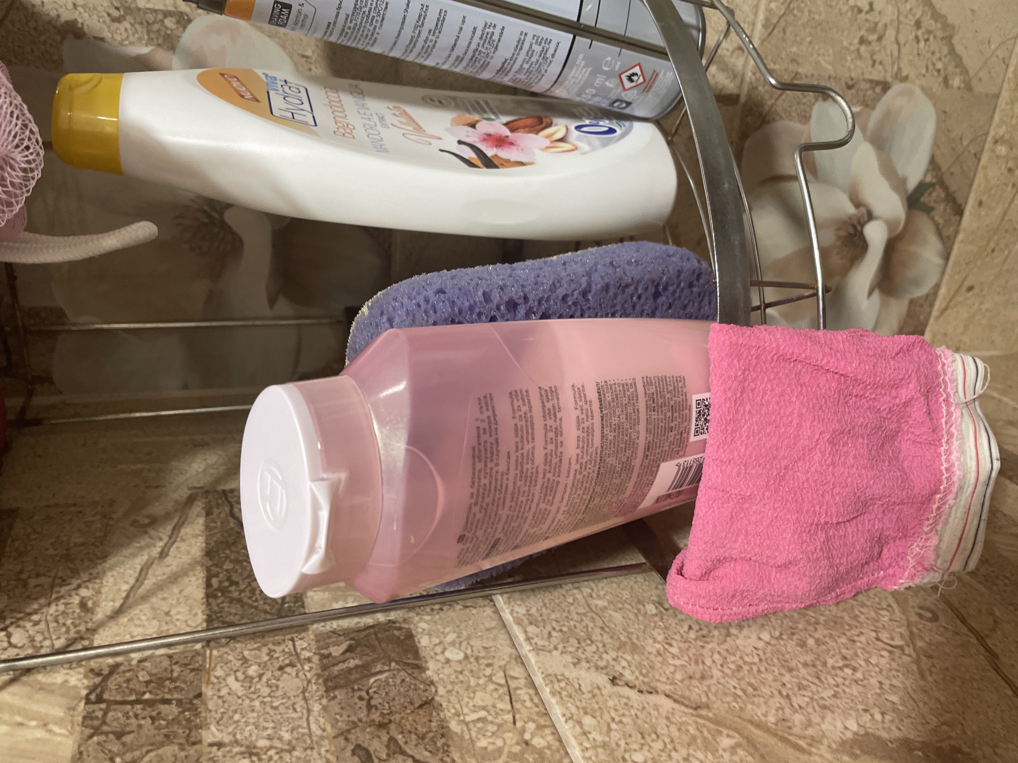 The image displays personal care products neatly arranged on a metal shower caddy in a bathroom setting in Ihtiman, Bulgaria. The background features light-colored wall tiles with a subtle floral pattern.
Prominently featured are two bottles:
1. A white bottle with a yellow flip-top cap, labeled "Hyolât Bagnodoccia MANDORLA E VANIGLIA EXTRACT," indicating an almond and vanilla bath foam.
2. A translucent pink bottle with a white flip-top cap. Its label contains extensive text in multiple languages, including phrases like "Formula obogaćena za 2x već sijaja" (formula enriched for 2x more shine) and "Sampon i balzam" (shampoo and conditioner), suggesting it is a hair care product. A QR code and the number "265113" are also visible on its label.
Other items include a purple sponge positioned beneath the pink bottle, and a pink scrubbing mitt or washcloth with white and red stripes on its edge, tucked beside the pink bottle. Partially visible at the top, behind the white bottle, is a silver spray can labeled "SHAVING FOAM Allantoin & menthol," with a "50 ml e" volume indicator and hazard symbols. Bulgarian text such as "улеснява бръсненето" (facilitates shaving) is also discernible on the spray can.
The items are stationary, suggesting they are simply stored in the shower area, ready for use. No specific activity is taking place, and there are no people present. The lighting suggests an indoor setting, but the time of day is not discernible.