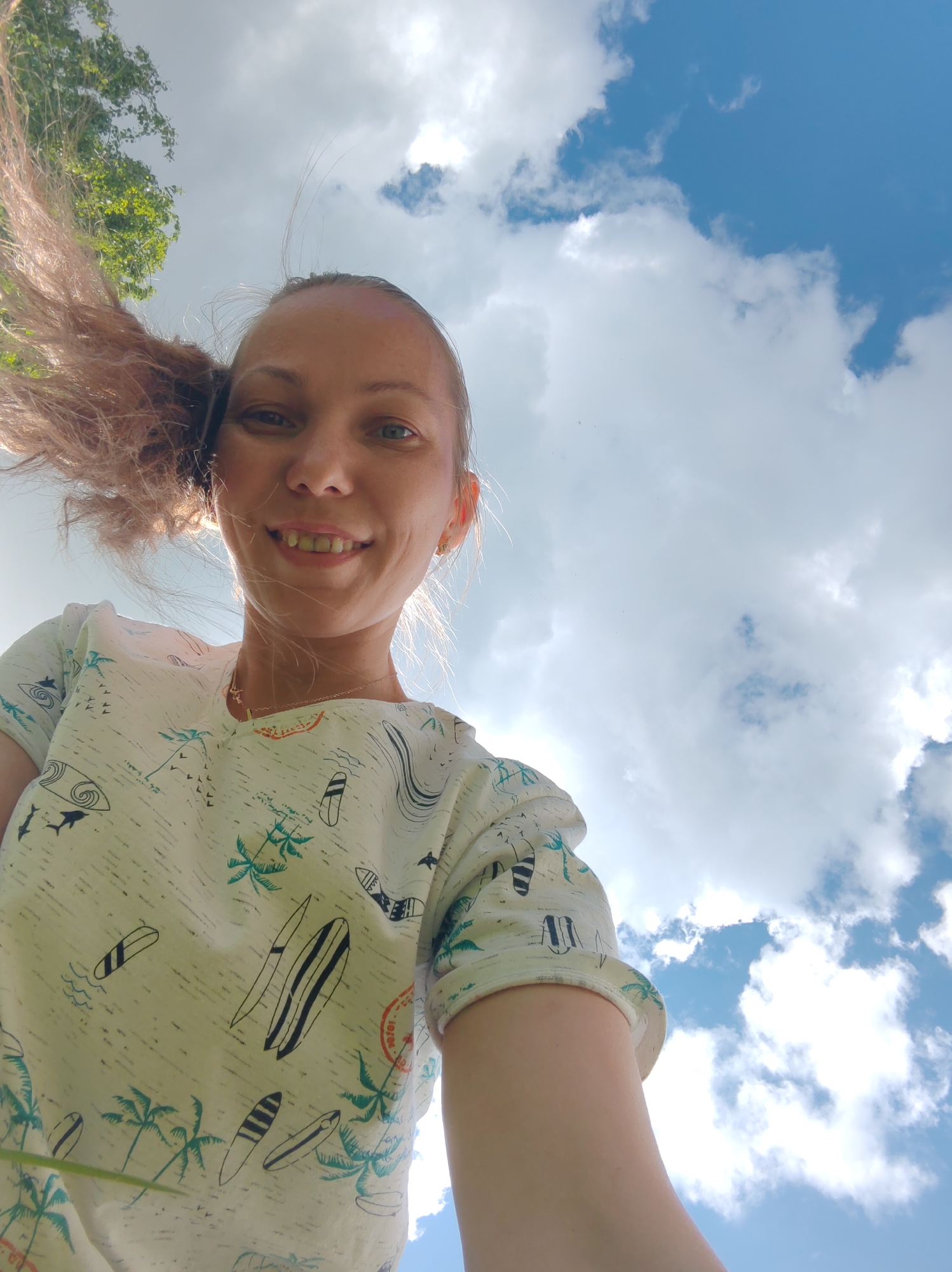 A low-angle, eye-level, medium shot captures a young adult woman from the chest up, looking directly at the camera with a smile. Her light brown hair is swept to her left by the wind. She is wearing a light-colored t-shirt with a repeating pattern of surfboards, palm trees, and waves. The background consists of a bright blue sky with scattered white clouds, and a glimpse of green foliage from a tree in the upper left corner. The image does not contain any elements that place it specifically in the city of Чухлэм, Russia.