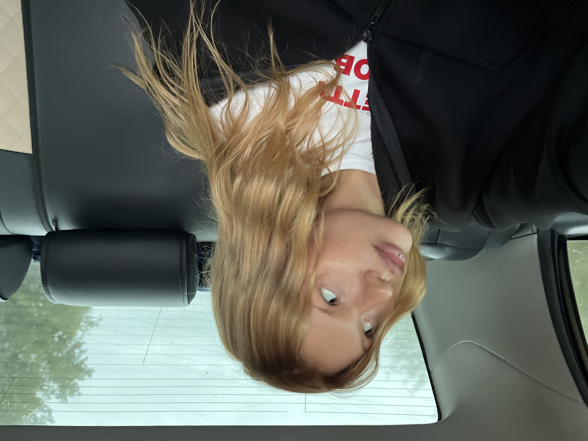 The image shows a young woman with blonde hair, wearing a white t-shirt with red lettering and a black jacket. She is positioned in the back seat of a car, with a headrest visible behind her. The car's rear window is in the foreground, and the outside appears to be overcast with trees visible through the glass. The photo is taken from an upside-down perspective. The visible text on her t-shirt reads "FETTO BOD."