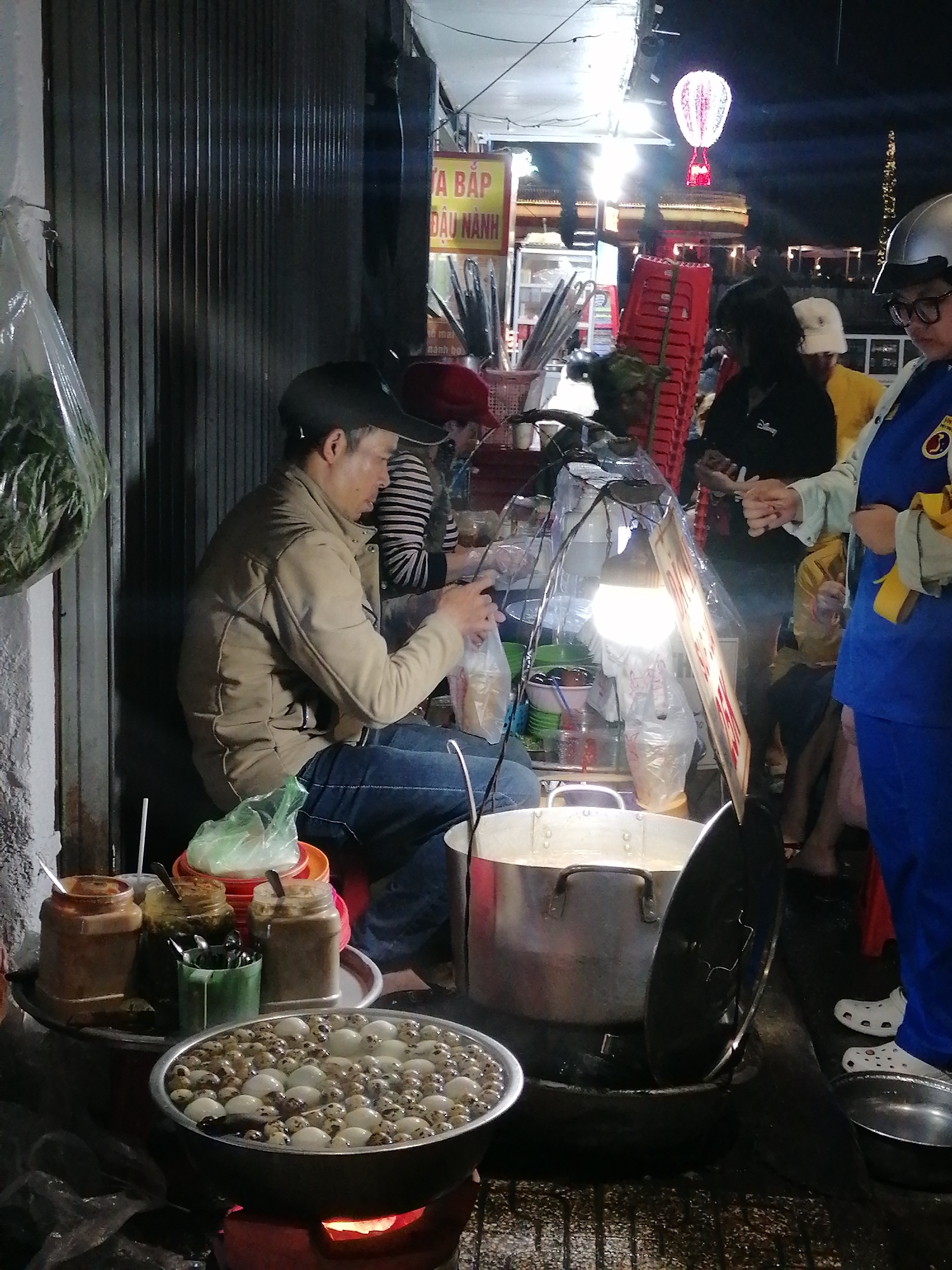 A street vendor prepares a food order at night in Da Lat, Vietnam. He sits next to a metal security gate. The vendor, wearing a tan jacket and baseball cap, fills a plastic bag. To his left is a low table with jars and a large bowl of small, white and brown speckled items, possibly quail eggs. Behind him, two people stand, one wearing a blue uniform and a silver helmet, handing money to another person. Behind them, red plastic chairs are stacked, and a sign with Vietnamese text is visible. Overhead, a string of lights illuminates the scene, with a hot air balloon-shaped light visible in the distance. A large pot sits on a burner near the vendor, and various kitchen items are scattered around the area. The environment appears to be a busy street at night.