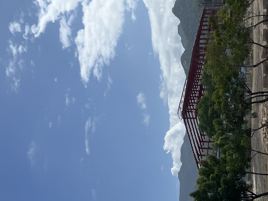 A blue sky with scattered white clouds dominates the scene. To the right, a red steel structure stands against a backdrop of mountains. Green trees partially obscure the structure. The ground, visible on the right edge, is a mix of dirt and rocks. The image is rotated, presenting the sky on the left.