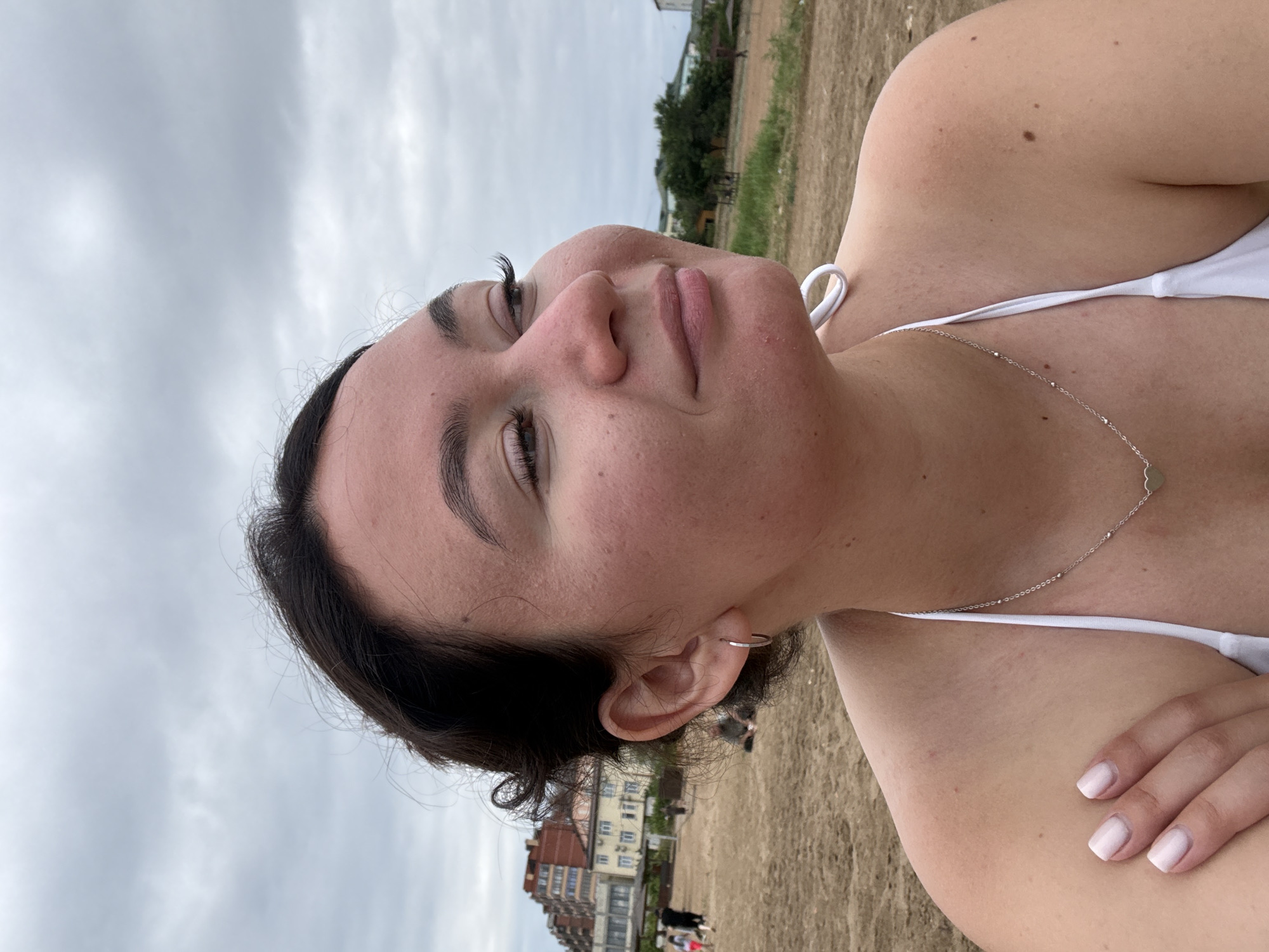 A young woman, with dark hair and fair skin, poses for a selfie on a beach. She is wearing a white bikini and a delicate silver necklace with a heart-shaped pendant. The camera is angled close to her face, capturing her eyes looking upwards and a slight smile. Her skin appears slightly sandy, hinting at the beach environment. In the background, the sand stretches out, leading to a body of water under a cloudy sky. Further in the distance, buildings can be seen, suggesting an urban area adjacent to the beach. The overall mood is relaxed and carefree, typical of a day at the seaside. The time of day appears to be daytime, with the overcast sky indicating a possibly cool or breezy day. The location is identified as Derbent, Russia, which is known for its coastline along the Caspian Sea. There is no visible text in the image.