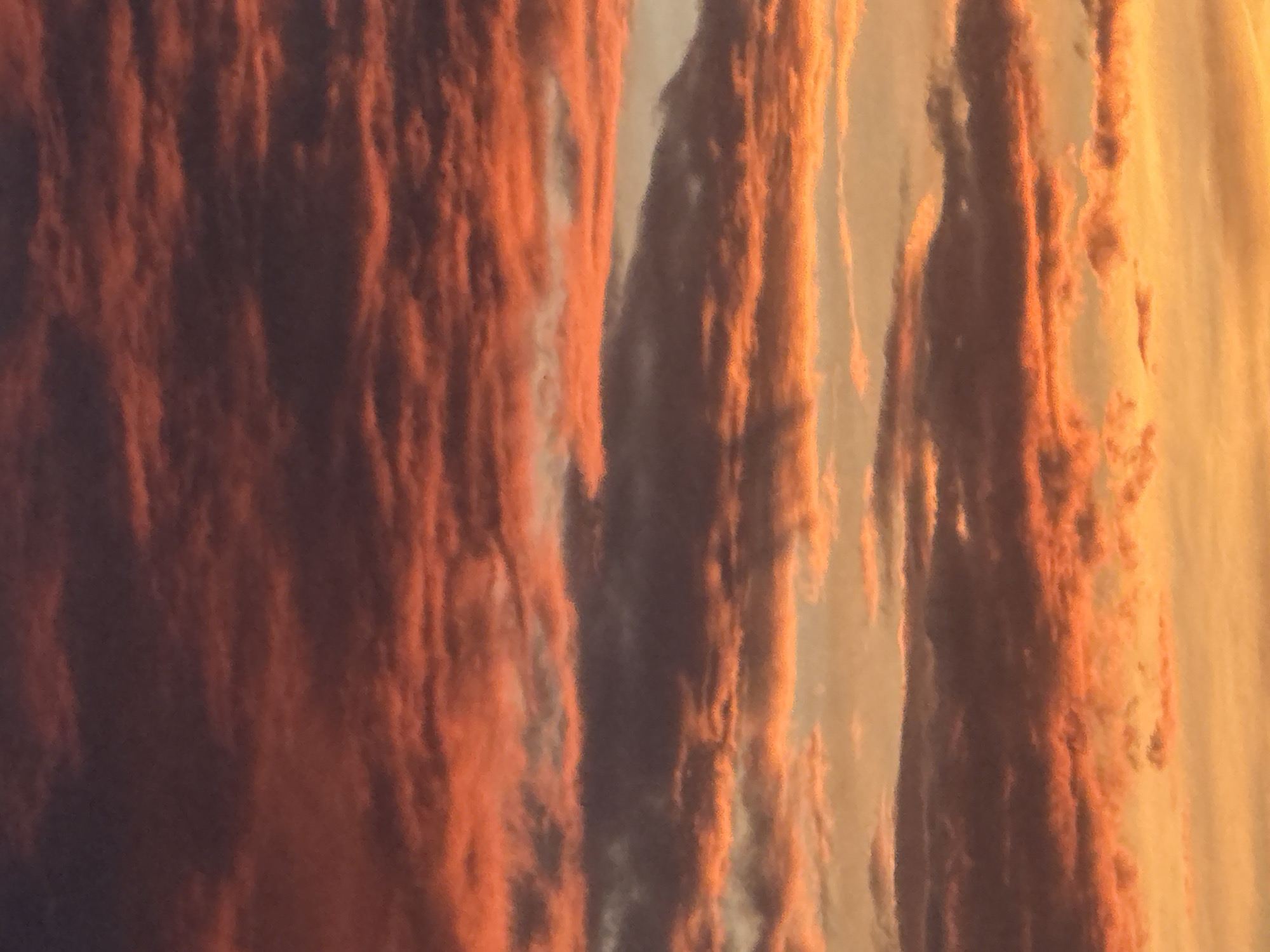 A photograph of the sky during sunset shows a dense layer of clouds, colored in hues of orange, red, and purple, with streaks of yellow peeking through the lower sections. The clouds are striated, appearing as if layered horizontally across the frame. The lighting suggests the sun is low on the horizon, casting a warm glow on the lower cloud formations and creating a gradient from dark purples at the top to bright yellows at the bottom. The image is a close-up view of the sky with no discernible features of a city or any other landscape elements.
