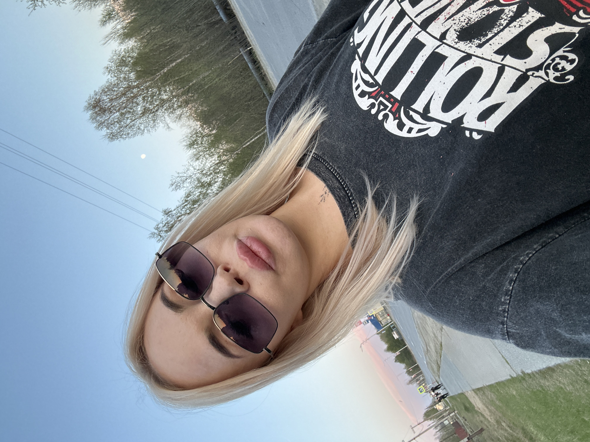 A woman with blonde hair and sunglasses is taking a selfie outdoors. She is wearing a black t-shirt with a white graphic design. The background includes a clear blue sky with a moon, trees, a concrete structure, grass, and part of a city street. The location is Высокий, Russia.