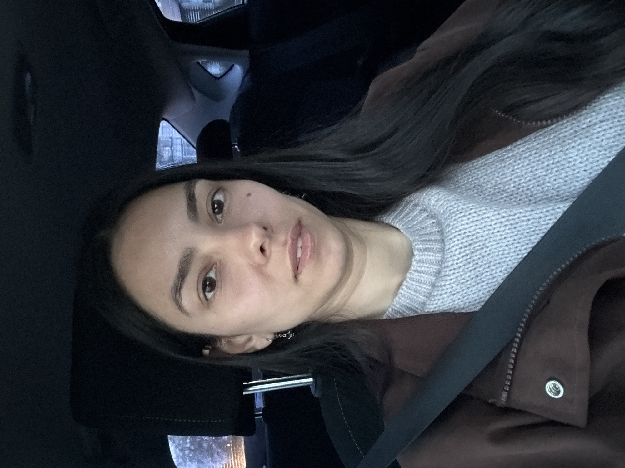 A portrait-style selfie, rotated 90 degrees counter-clockwise, features a woman seated inside what appears to be a car. The woman, positioned on the left side of the frame, looks directly at the viewer with a neutral expression and slightly parted lips. She has long, dark brown hair that falls over her right shoulder (viewer's left). Her eyes are dark brown, and a small mole is visible on her right cheek. She is wearing a light grey knitted sweater under a dark brown jacket, which has a visible zipper and a metallic eyelet detail. A black seatbelt diagonally crosses her chest and jacket from the upper right to the lower left of the frame. The background reveals the dark interior of the car, with blurred lights and shapes visible through the windows, suggesting an evening or nighttime setting. The overall lighting is dim. No text is visible in the image.