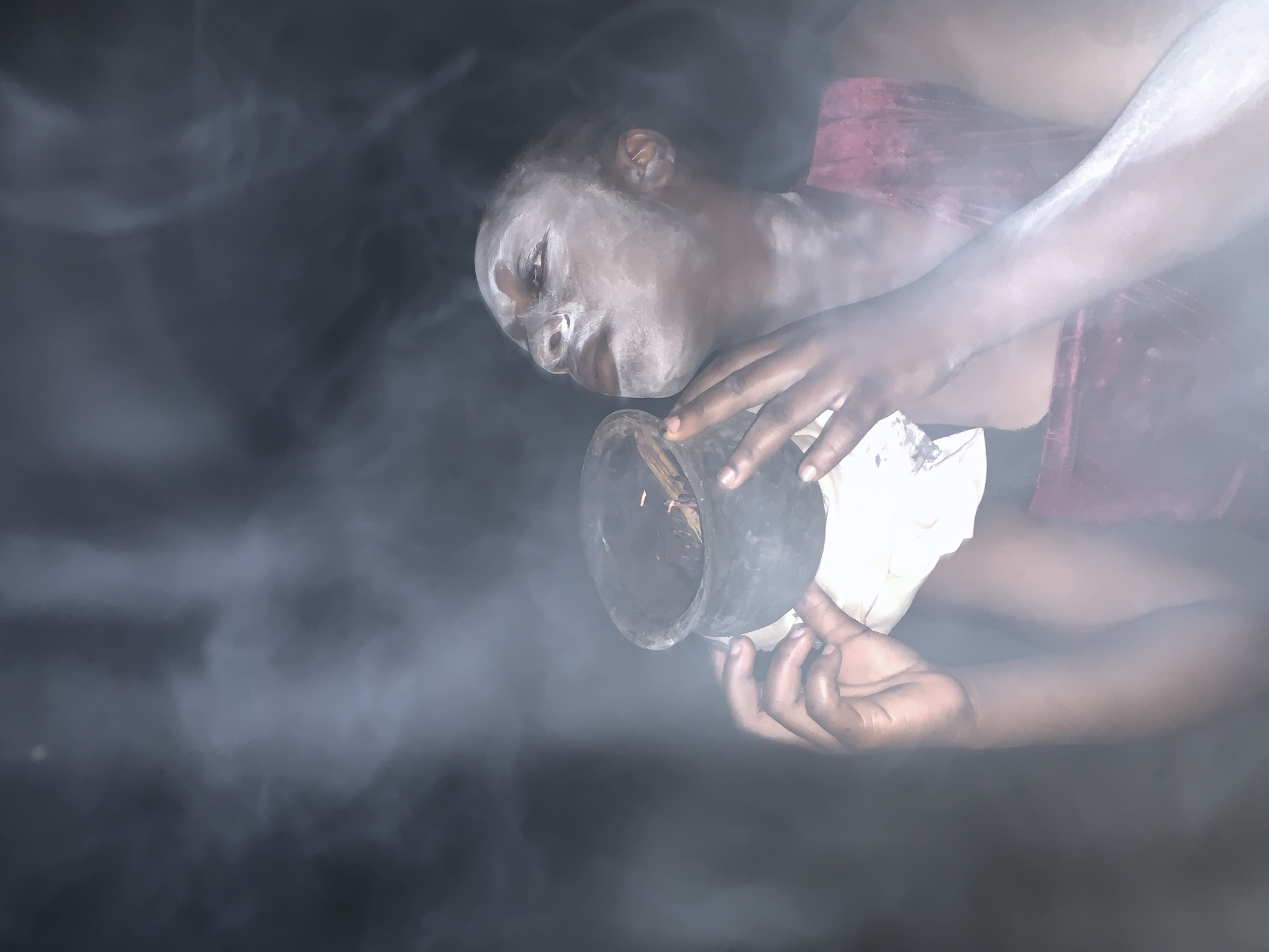 In a poorly lit environment, a person with white markings on their face and wearing a reddish garment is holding a dark container with lit contents, possibly incense, emitting smoke. The setting is implied to be associated with Agban, Nigeria. The person is looking down at the container.