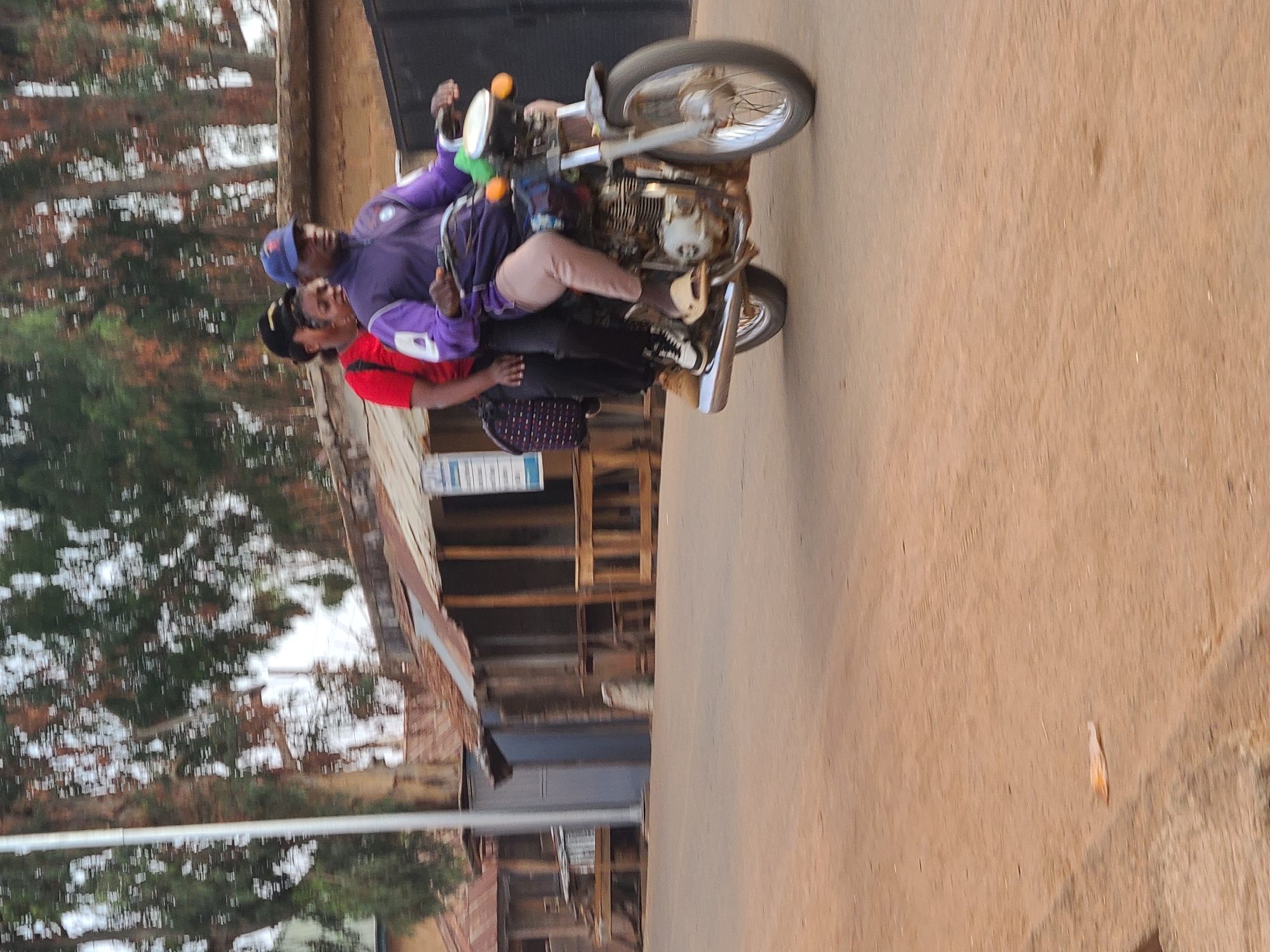 The image shows two people riding a motorcycle on a road, likely within an urban area. The motorcycle appears to be an older model. The driver is wearing a blue cap and a purple shirt, and the passenger is wearing a red shirt. They are both wearing pants. The environment seems to be a street in a city. The location is unknown, and that of Jos, Nigeria cannot be confirmed.