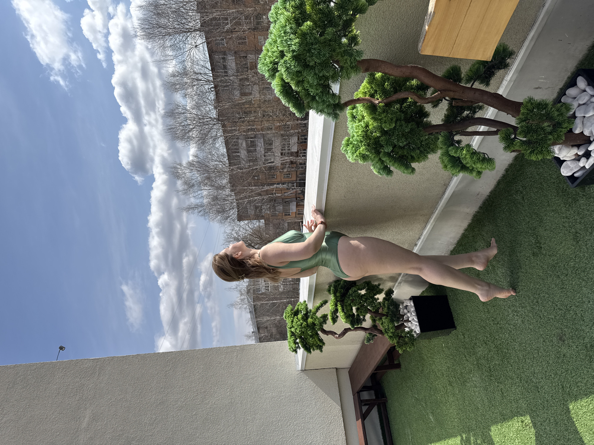 A woman wearing a green swimsuit is standing on a balcony in Votkinsk, Russia. She is leaning on the edge of the balcony, looking up at the sky, which is partially cloudy. In the background, there is an apartment building and trees with bare branches. A potted bonsai tree sits on the balcony. The floor of the balcony is covered in artificial grass.