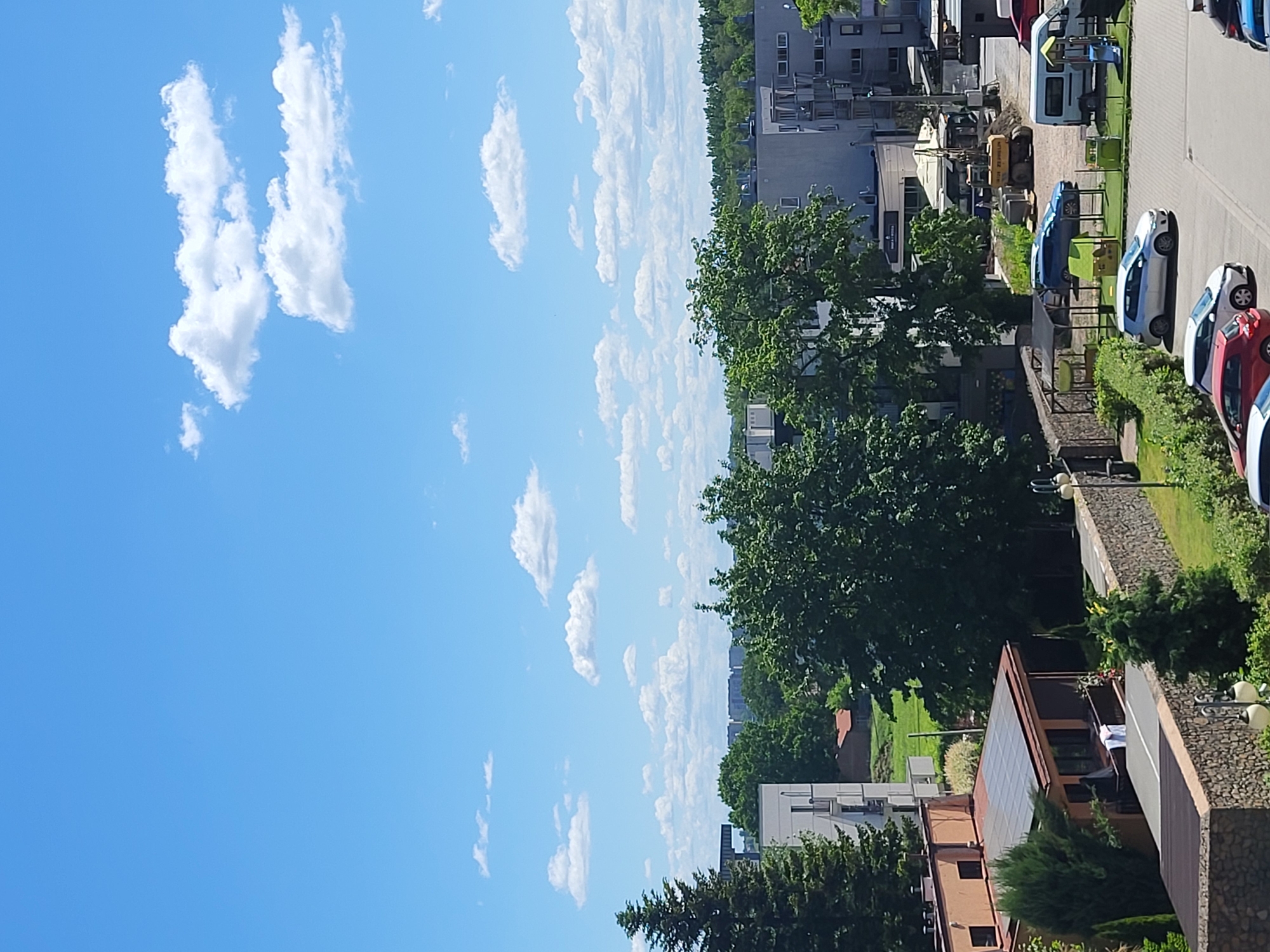 The image shows a cityscape under a bright blue sky with scattered cumulus clouds. Buildings of varying heights and architectural styles are visible, interspersed with green trees. A parking lot is partially visible, with multiple cars parked in a row. The photograph appears to have been taken from an elevated vantage point.