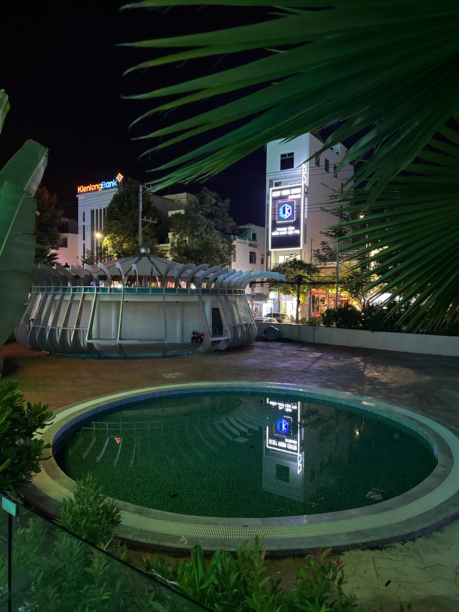At night in Nha Trang, Vietnam, a circular swimming pool reflects the neon signage of a modern building. The pool's surface is dark green, with submerged lights creating white patterns. In the background, a building displays illuminated text, including "KienlongBank" in red and yellow, and another sign with a blue hexagonal logo and white text that reads "REX AERO CENTER." A unique, open-air, futuristic-looking structure with a domed roof and metal framework stands between the pool and the building. Lush green foliage from palm trees partially obscures the view.