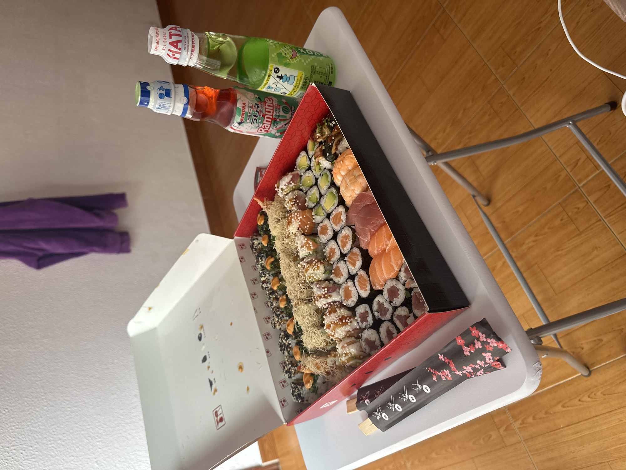 An overhead shot shows a large takeout box of sushi and two bottles of Ramune soda sitting on a white folding table. The sushi box is red and black and open, revealing multiple rows of sushi. From left to right, the rows appear to contain salmon, tuna, avocado, shrimp, eel, and various rolls covered in sesame seeds and other toppings. The Ramune bottles are glass with colorful labels. The red bottle contains a red liquid, and the green bottle contains a green liquid. The table is set up on a wooden floor. In the background, a purple garment hangs on a white wall. The scene is indoors, likely in a residential setting. The specific location in Oviedo, Spain, is not identifiable from the image.