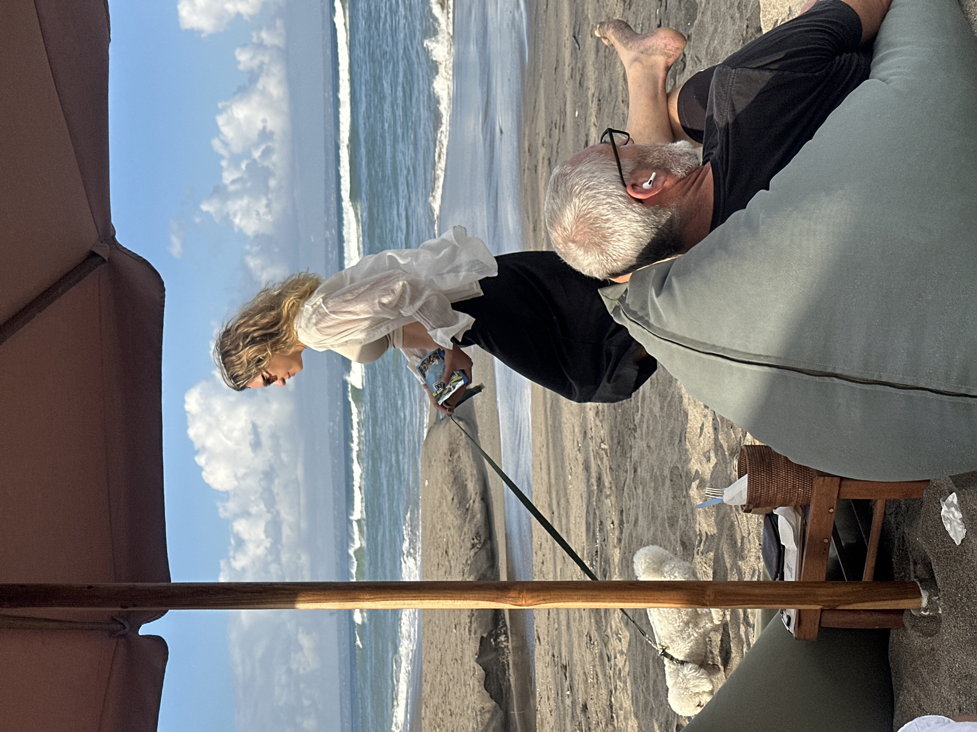A woman with blonde, wavy hair and sunglasses walks her white poodle on a leash across a sandy beach in North Kuta, Indonesia. The woman is wearing a sheer white long-sleeved shirt over a light-colored top and dark pants. She is holding a magazine in her left hand. In the foreground, a man with gray hair and glasses is reclining on a large, light green beanbag chair, facing away from the camera. He is wearing a black t-shirt and appears to have earbuds in his ears. A small wooden table with a woven basket containing utensils and napkins sits between the man and the woman's dog. The ocean with waves is visible in the background, under a blue sky with scattered white clouds. A brown beach umbrella shades a portion of the scene.