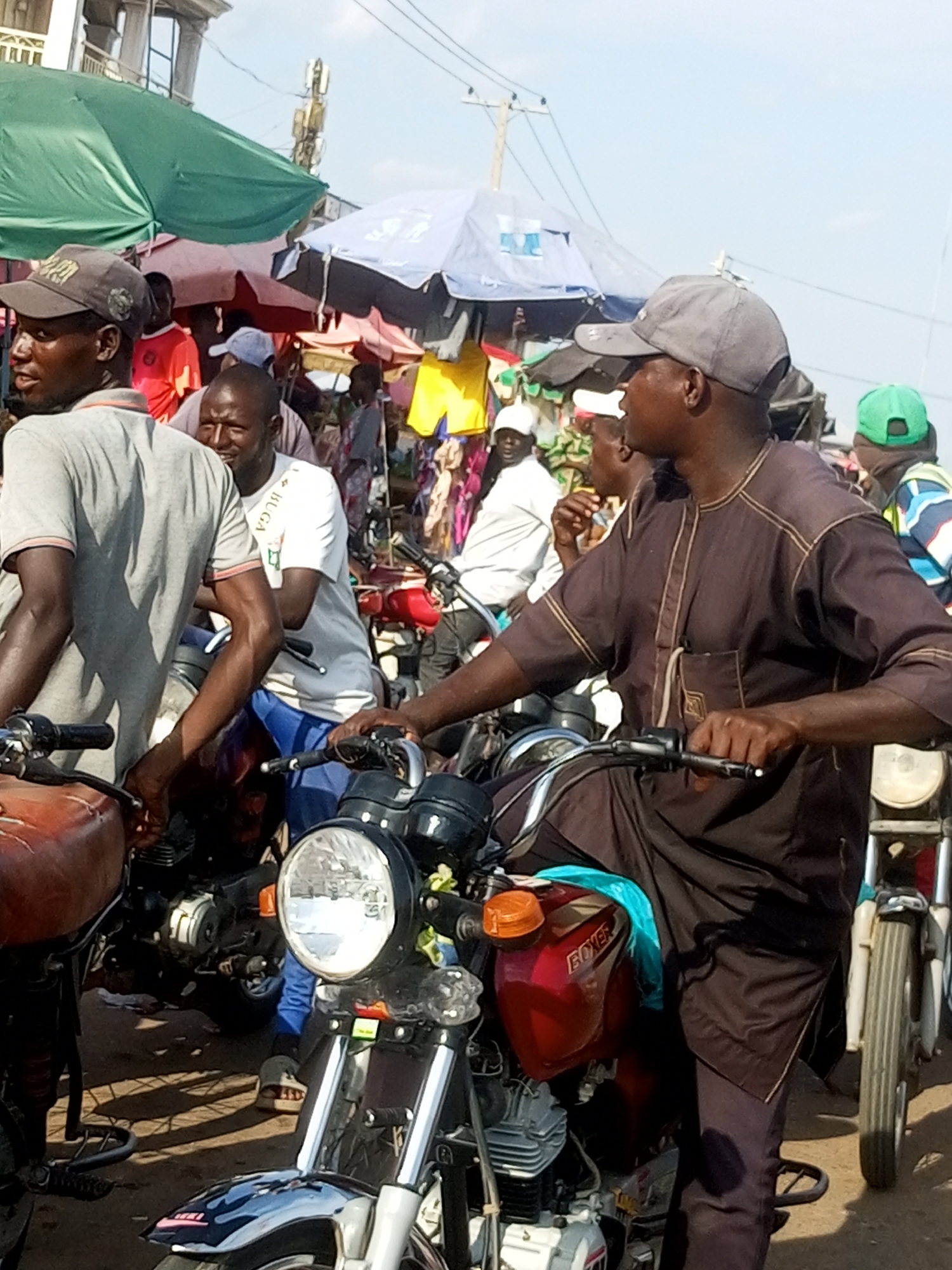 A busy marketplace scene is depicted in Gombe, Nigeria, with several men on and around motorcycles. The foreground is dominated by a motorcycle with a bright red fuel tank that has "BOXER" written on it in white lettering. A person wearing a dark brown tunic and dark pants is seated on this motorcycle, holding the handlebars. To the left, another motorcycle with an orange fuel tank is visible.
Behind the foreground motorcycles, more people are seen, many wearing caps. One man in a grey t-shirt and a cap is standing near the motorcycles, facing left. Another man in a white t-shirt and blue shorts is also near the motorcycles. Further in the background, the market stalls are apparent, with various umbrellas providing shade, including green and blue ones. Some stalls display colorful clothing. The overall environment suggests a crowded and active outdoor market setting. The lighting indicates it is daytime.