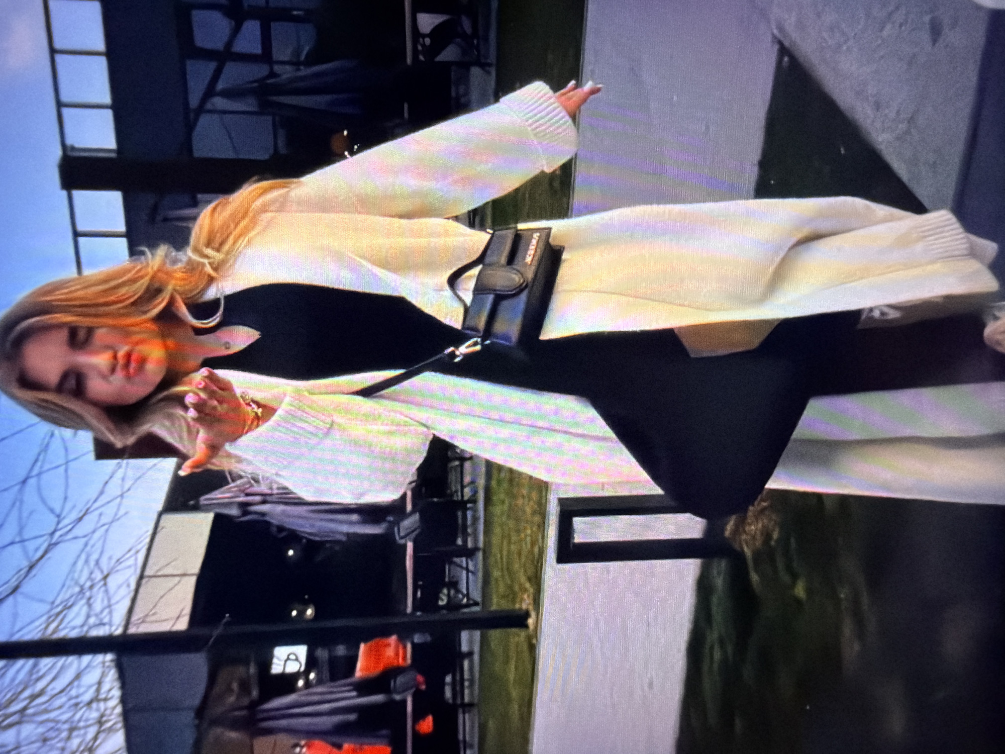 A young woman with blonde hair is standing outdoors in a city setting, possibly in Beșalma, Moldova, based on the context provided. She is wearing a long, cream-colored cardigan over a black dress and is carrying a small black handbag. The woman is leaning forward, puckering her lips as if blowing a kiss, and has one hand extended towards the camera. The background shows outdoor seating areas with tables and chairs, suggesting a cafe or restaurant. The lighting indicates it might be dusk or early evening.