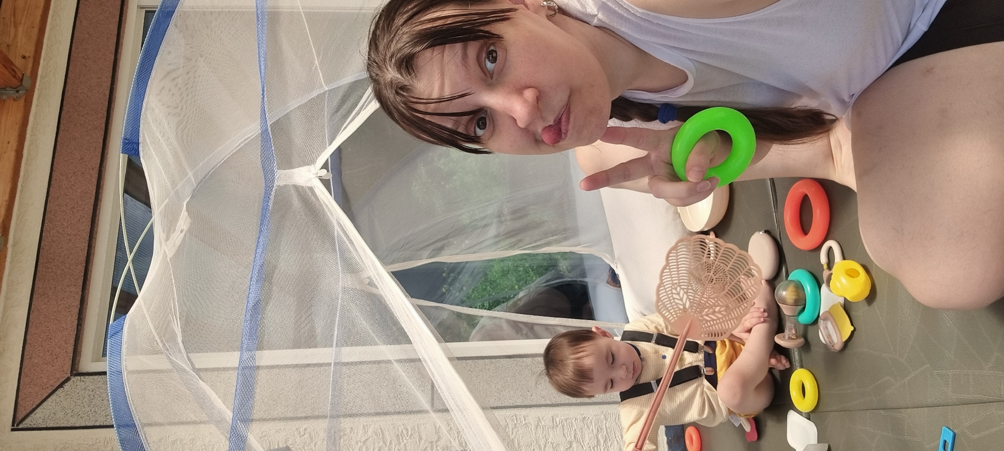 The image shows a woman and a baby inside what appears to be a portable mosquito net tent. The woman is holding a green plastic ring and making a peace sign with her hand, while the baby is playing with toys on the floor. The toys include plastic rings, a rattle, and a fly swatter. The environment is indoor. Given the information provided, the location is Dunnae-myeon, South Korea.