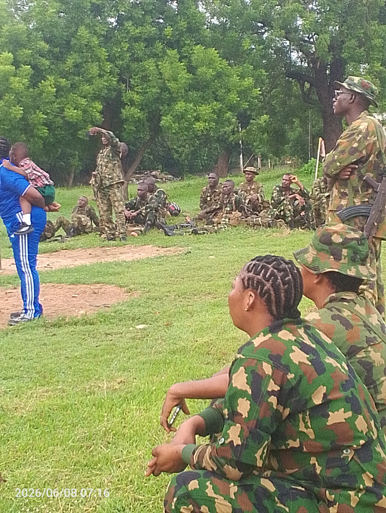 In Adankolo, Nigeria, a group of soldiers are gathered outdoors on a grassy field, with trees in the background. A man in a blue tracksuit carries a child on his left shoulder, pointing towards the right. Several other soldiers in camouflage uniforms are seated or standing in the middle ground and background. Two women in camouflage uniforms are in the foreground, facing away from the camera. The environment appears to be a training or gathering area.
