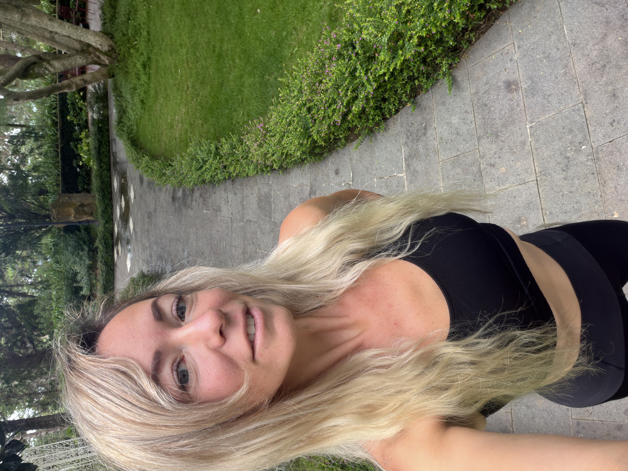 A blonde woman in a black sports bra and black leggings takes a selfie in a park with a paved walkway lined by manicured hedges. A grassy area with trees and some red chairs is visible to the right of the walkway. The walkway has a damp appearance, suggesting recent rain. The woman is smiling and looking at the camera.