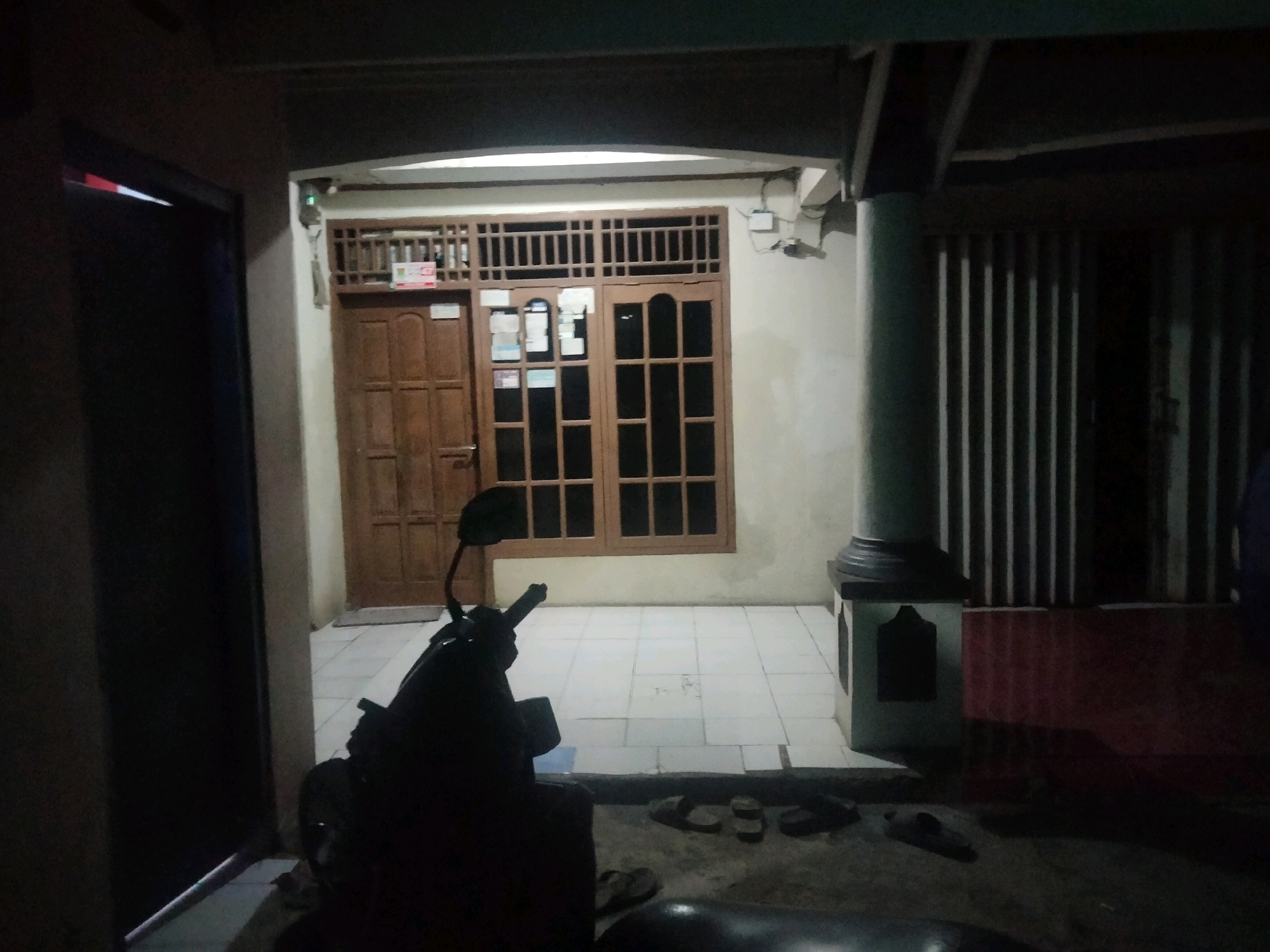 The image depicts an outdoor area at night, likely a front porch or entryway, in Suka Sari, Indonesia. In the foreground, a dark-colored motorcycle is parked, silhouetted against the dim lighting. To the left, a dark doorway or entrance is visible. Beyond the motorcycle, a tiled floor leads to a wooden door and a large, multi-paned window with wooden frames. The window has a decorative wooden grill at the top. Signs are affixed to both the door and the window. A concrete pillar, painted a light color and topped with a dark cap, stands to the right of the window. Behind the pillar, a gated or barred structure is present, with vertical elements casting shadows. Several pairs of dark-colored sandals are scattered on the ground in front of the tiled area. The overall lighting is dim, suggesting it is nighttime, with artificial light sources illuminating the immediate vicinity.