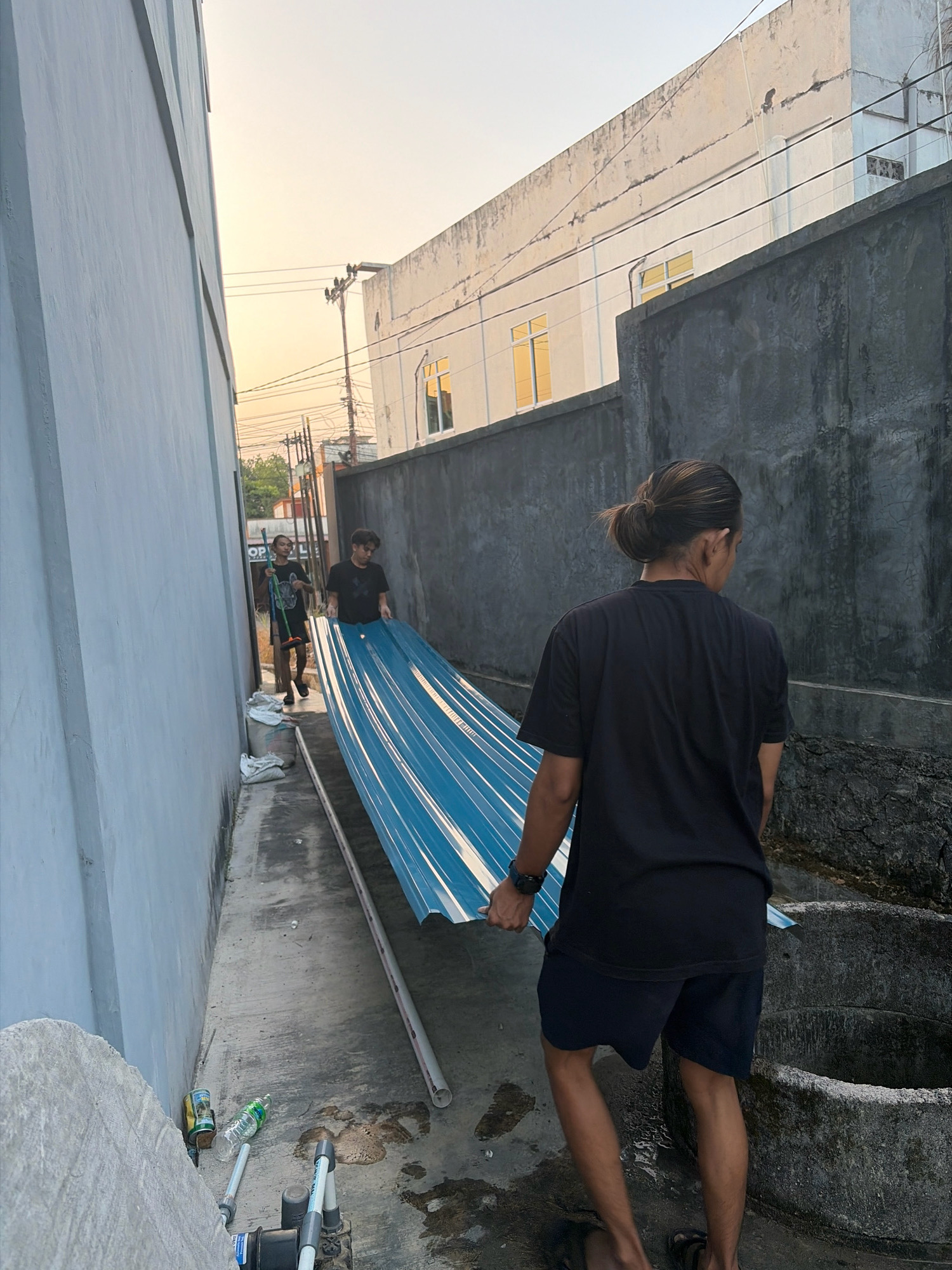 In Tanjung Pinang, Indonesia, two young men are seen carrying a long, blue corrugated metal sheet down a narrow alleyway. The man in the foreground, with his back to the camera, has his hair tied in a bun and wears a black t-shirt and dark shorts. He is holding the metal sheet with his left hand. Behind him, another man in a black t-shirt and dark shorts, is walking and also holding the metal sheet. Further down the alley, a third individual, partially obscured, is seen carrying a broom. The alley is flanked by two buildings; a light-colored one on the left and a dark, rough-textured wall on the right. The ground is concrete and appears damp in places, with some debris, including a plastic bottle and a can, scattered near the building on the left. Overhead, power lines are visible against a bright, hazy sky, suggesting it is either sunrise or sunset. A large, circular, concrete structure, possibly a well or drain, is visible in the lower right corner of the image.