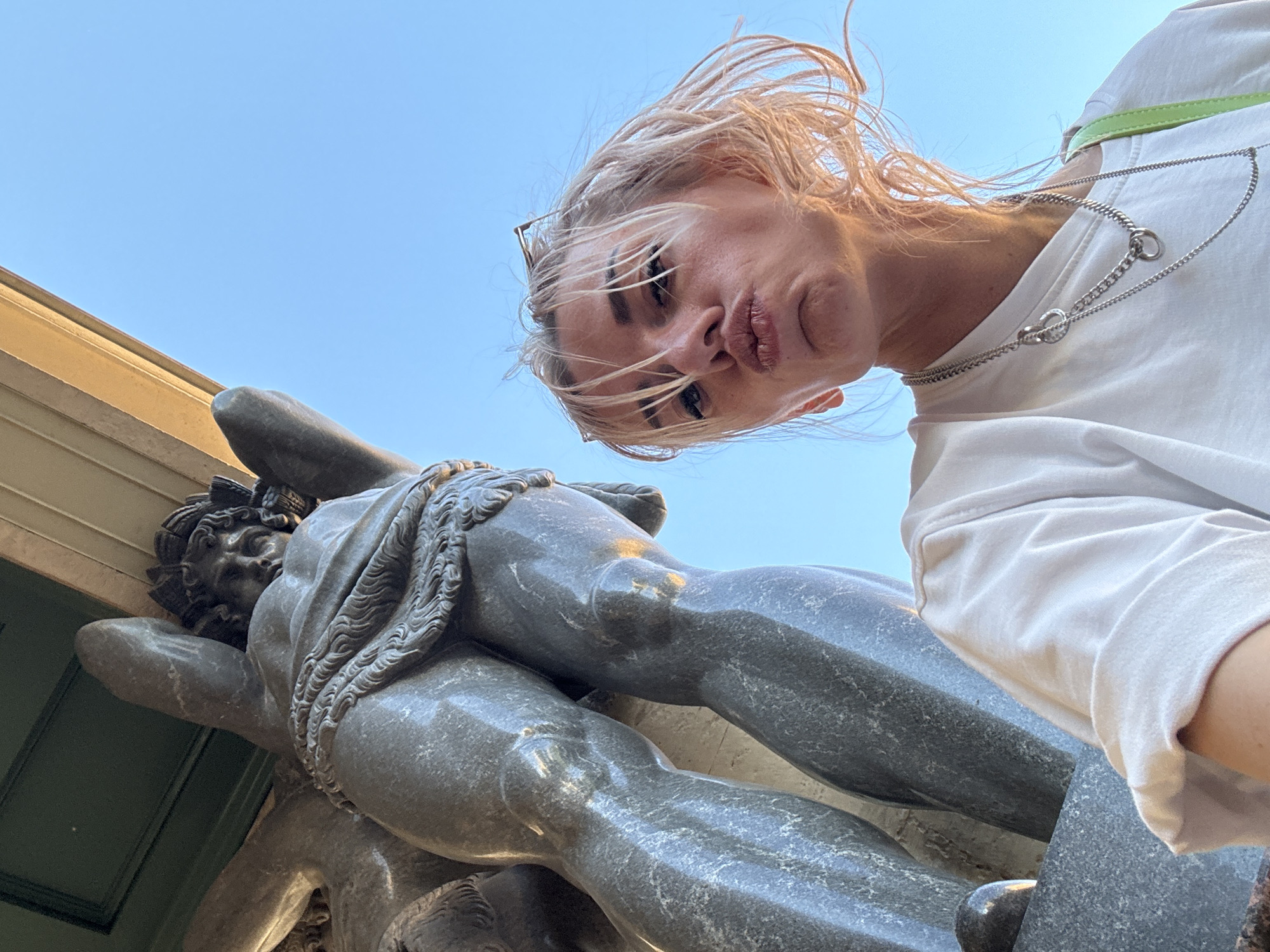 The media file is a low-angle, close-up photograph featuring a young woman with blonde hair and a white t-shirt, posing for a selfie. She is looking directly at the camera with a playful expression, her lips puckered. Her hair is windswept, suggesting an outdoor setting with a breeze. She is wearing a silver necklace with circular links.
In the foreground, partially obscuring the woman, is a large, dark grey stone statue of a muscular male figure. The statue appears to be part of a larger monument or sculpture. The woman is positioned above and slightly behind the statue, creating a dynamic perspective. The statue's torso, arms, and legs are visible, rendered in a classical style with intricate details like musculature and drapery.
The background is a clear, bright blue sky, indicating a sunny day. A sliver of a tan-colored building facade is visible in the upper left corner, providing a hint of the surrounding architecture. The lighting suggests it is daytime, likely late afternoon or early evening given the angle of the light. The overall scene conveys a sense of playful interaction between the woman and the historical statue in a public outdoor space, possibly a park or square in Saint Petersburg, Russia, given the location context. There is no visible text in the image.