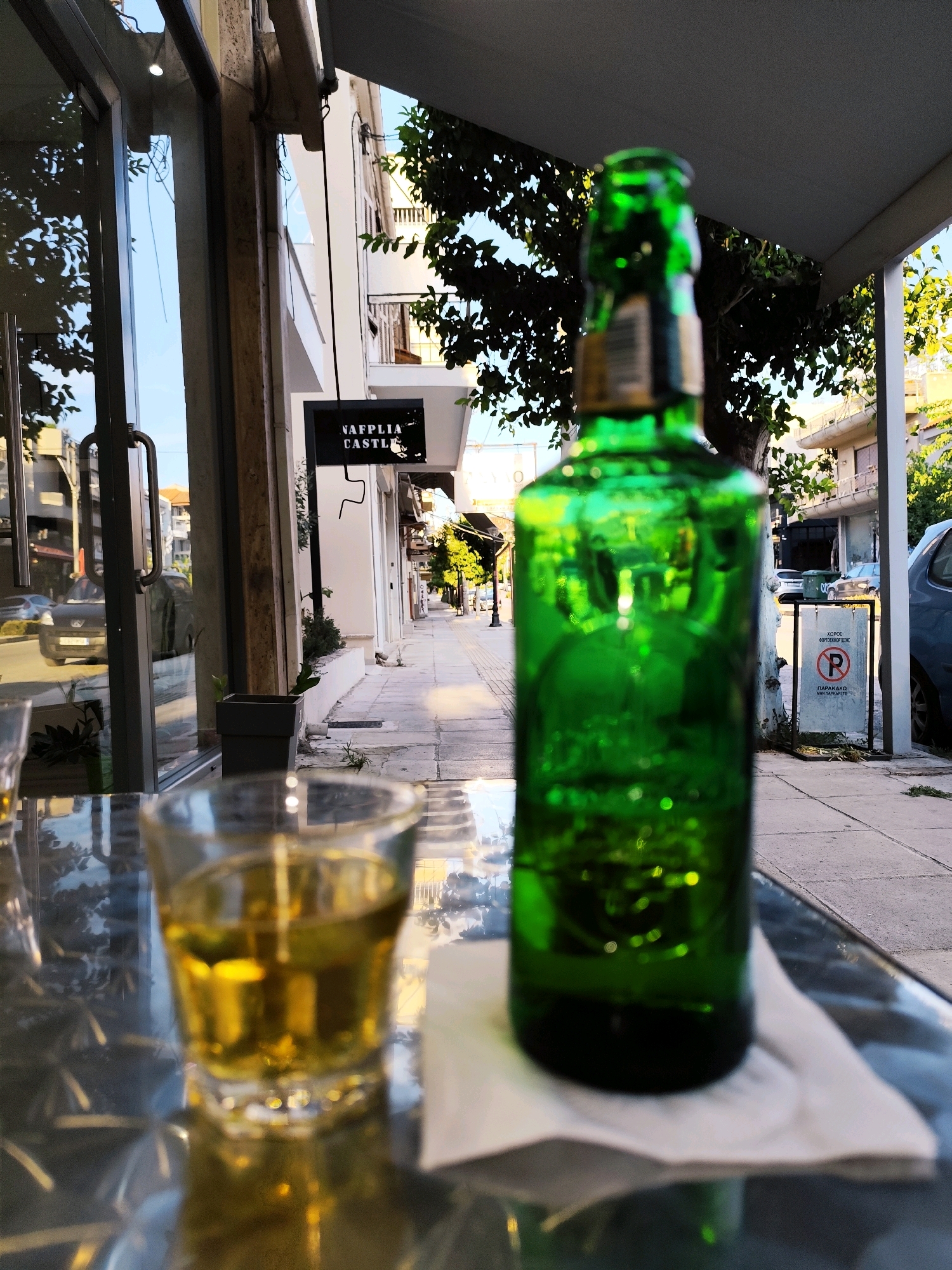 A low-angle, close-up shot captures a green glass bottle of beer and a shot glass filled with a golden liquid. The scene is set outdoors on a glass-topped table. In the background, a street in the Municipal Unit of Nafplio, Greece, is visible, with buildings and trees lining the sidewalk. A sign with the words "NAFPLIA CASTLE" is mounted on a building. A parking restriction sign, featuring a red circle with a diagonal line through a "P," is also present. The lighting suggests it is late afternoon or early evening.
