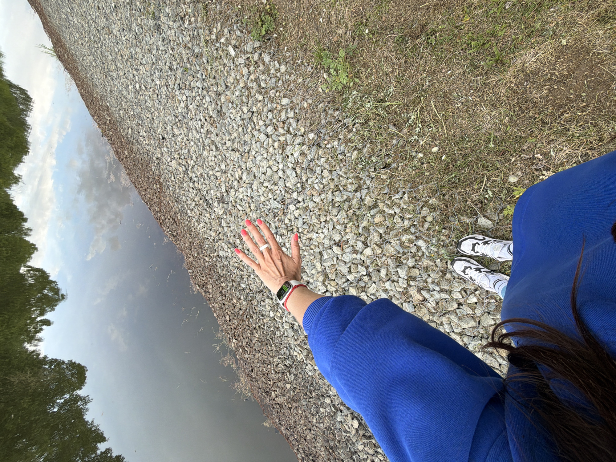 A first-person, high-angle shot captures a person standing by a body of water, likely a canal or river, in Birobidzhan, Russia. The individual is wearing a vibrant royal blue long-sleeved top or sweatshirt, paired with white and black sneakers and white socks. Their dark hair cascades over their right shoulder. The person's left hand is extended forward, featuring bright red or coral painted fingernails, a white ring on the ring finger, and a white digital watch with a red band on the wrist.
The ground immediately around the person consists of a bank covered in light grey and brown gravel, transitioning into dry grass and sparse weeds further back. To the left, a calm expanse of water reflects the sky, showing light blue patches with clouds, and the dark, inverted images of trees that appear to line the opposite bank. The reflections suggest a bright, possibly overcast, daytime. The activity is seemingly a moment of observation or posing by the water's edge. No discernible text is visible on the watch or other items.