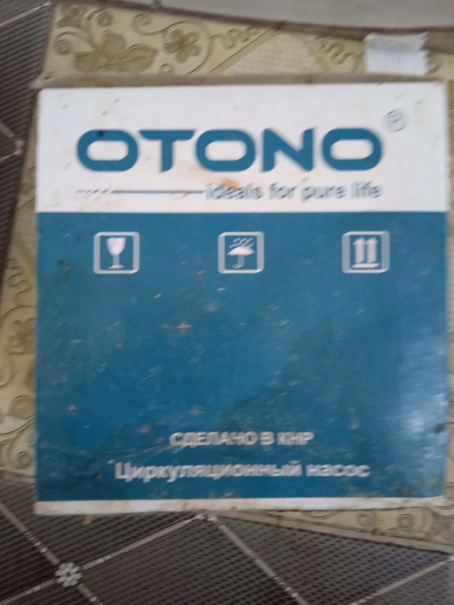 The image shows a close-up of a blue and white product box. The top portion of the box features the brand name "OTONO" in large blue letters, with a smaller tagline below that reads "ideals for pure life". There are three universal shipping symbols below the tagline: a wine glass inside a square, an umbrella inside a square, and the Roman numerals "II" within a square. The bottom portion of the box is solid blue and contains text in Russian. The text reads "СДЕЛАНО В КНР" which translates to "MADE IN PRC" and below that "Циркуляционный насос" which translates to "Circulating pump". The box is slightly dusty and appears to be on a patterned surface that resembles a floor. The lighting is overhead and casts a slight shadow on the left side of the box.