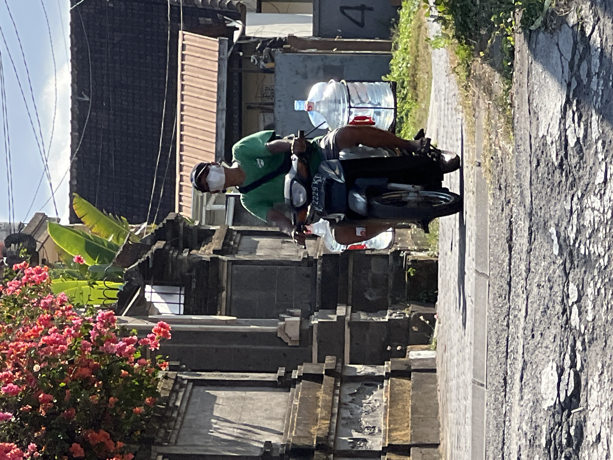 The image depicts a person riding a motorcycle in Denpasar, Indonesia, on a sunny day. The rider, wearing a green t-shirt, dark shorts, a dark baseball cap, and a white face mask, is operating a dark-colored motorcycle. Attached to the rear of the motorcycle are two large, clear plastic water jugs. The motorcycle's license plate is partially visible, showing the characters "2229 KT".
The motorcycle is on a paved road that appears worn and cracked in some areas, with a distinct curb separating it from a sidewalk made of interlocking stones. To the left of the road, a textured gray stone wall with decorative elements and steps is visible. Above the wall, a vibrant bush with clusters of pink flowers and lush green leaves hangs into the frame. In the background, several buildings with dark tiled roofs and some with reddish-brown corrugated metal roofing are present. Some of these structures appear to be residential or commercial. Green foliage and vegetation are abundant along the roadside and in the background, suggesting a tropical environment. The sky above is clear and blue with a few faint white clouds.