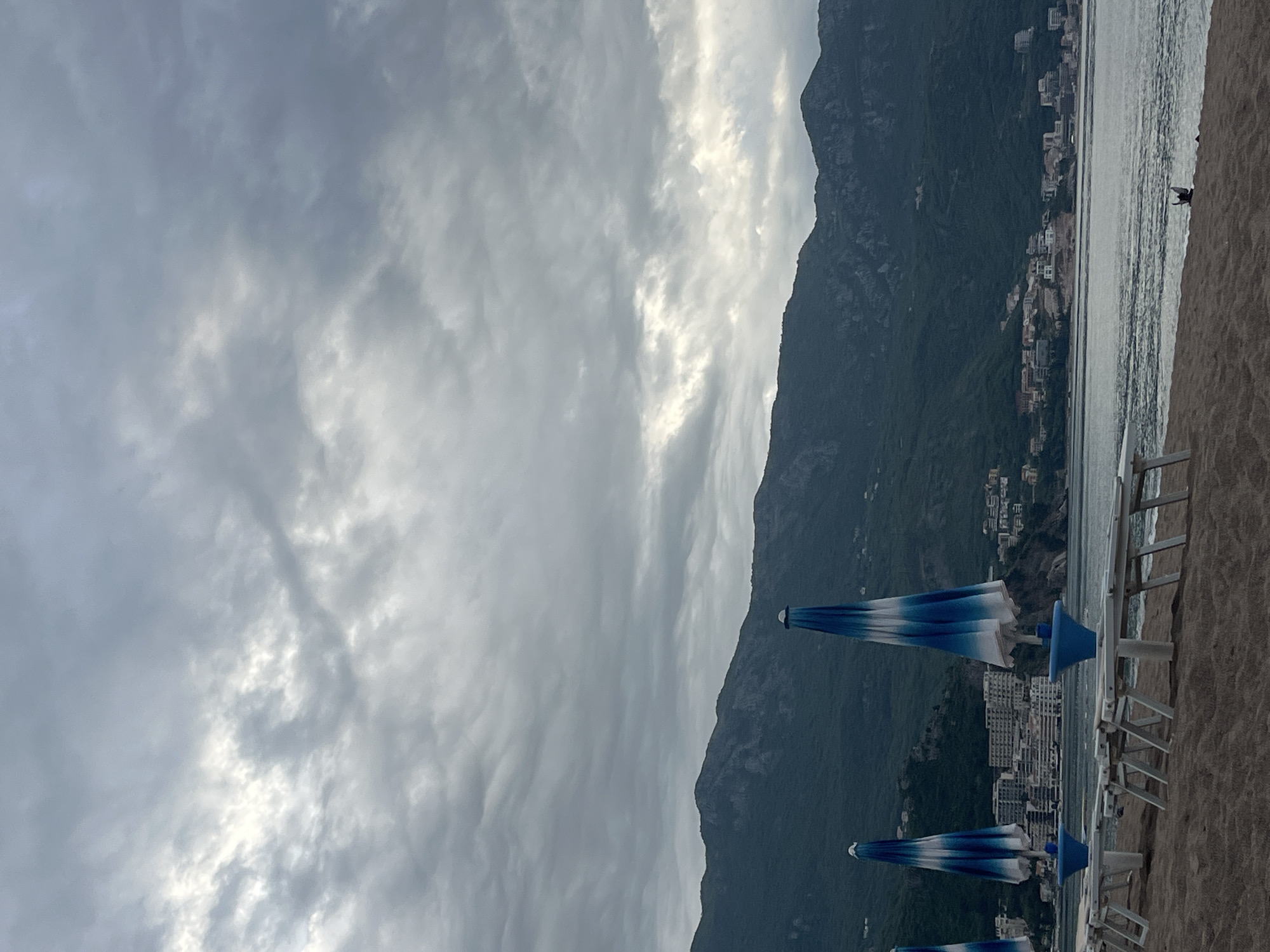 The image shows a beach scene in Boreti, Montenegro, under an overcast sky. A mountain range runs along the center of the frame, with buildings nestled at its base and along the coastline. The beach is to the right, with a row of white beach chairs and blue and white striped umbrellas. The water is visible as a thin strip between the beach and the buildings. A silhouette of a person is visible at the far right on the beach.