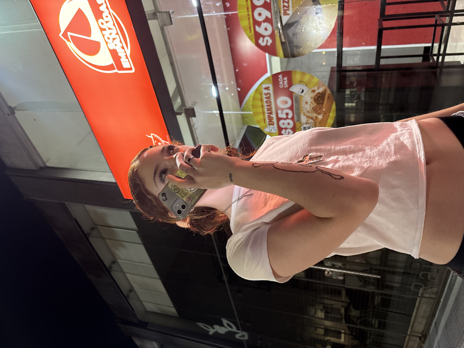 A fair-skinned woman with reddish-brown hair stands outside a restaurant, holding a phone to her ear. She wears a white crop top and a silver necklace. An abstract dark tattoo is visible on her right arm. Behind her, the restaurant's signage includes text in Spanish and prices for pizza and empanadas. A glass window reflects additional details of the restaurant interior, including what appears to be a red wall and black shelving.
