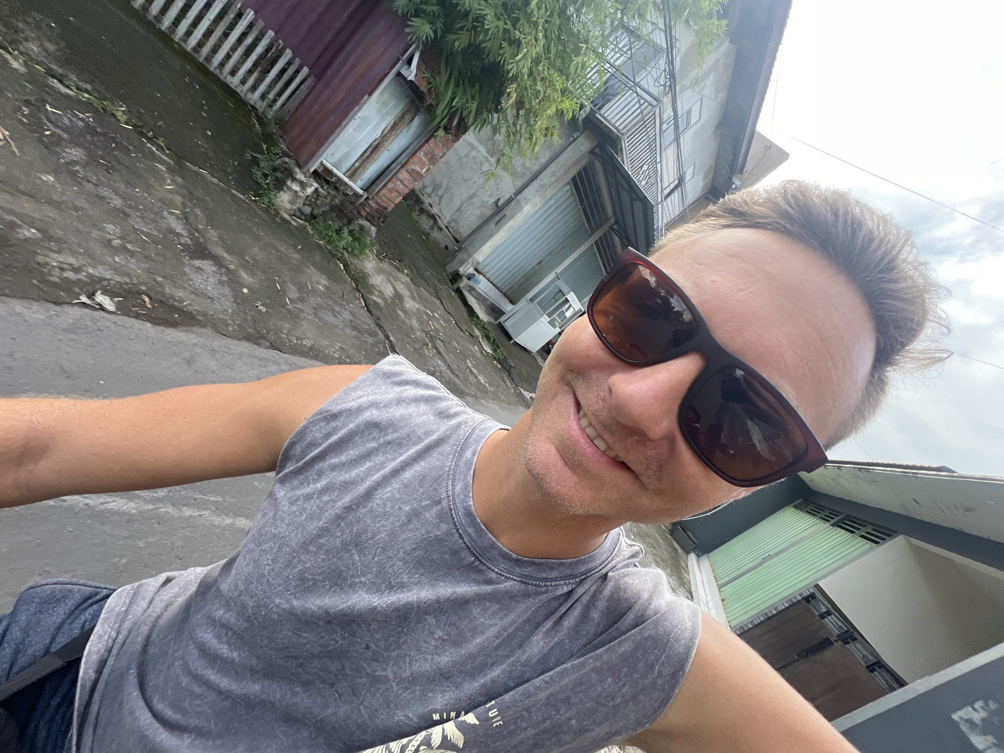 A medium-shot selfie taken from a low angle shows a man wearing sunglasses and a grey t-shirt. He is smiling slightly. The background includes a street scene with buildings. The location is identified as Sukawati, Indonesia based on visual elements of buildings and architecture. The buildings in the background appear to be constructed from concrete and corrugated metal. The street surface is cracked and uneven.