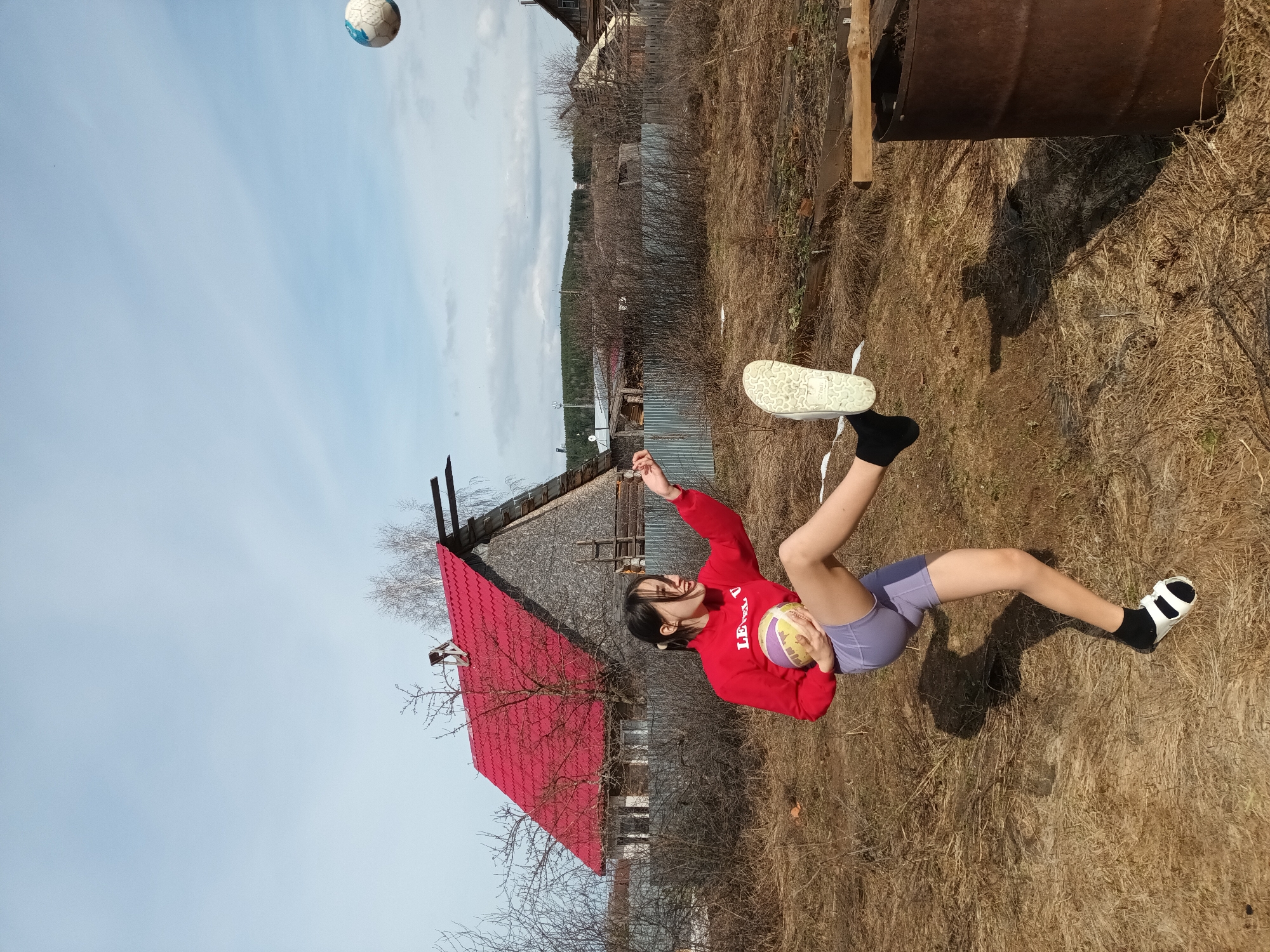 A young woman is outside, kicking a soccer ball into the air. She's wearing a red sweatshirt, purple shorts, black socks, and sandals. She is holding another ball under her arm. The scene is in a rural area with a red-roofed house and dry grass. A metal barrel is also visible. The location is likely in Ольховка, Russia.