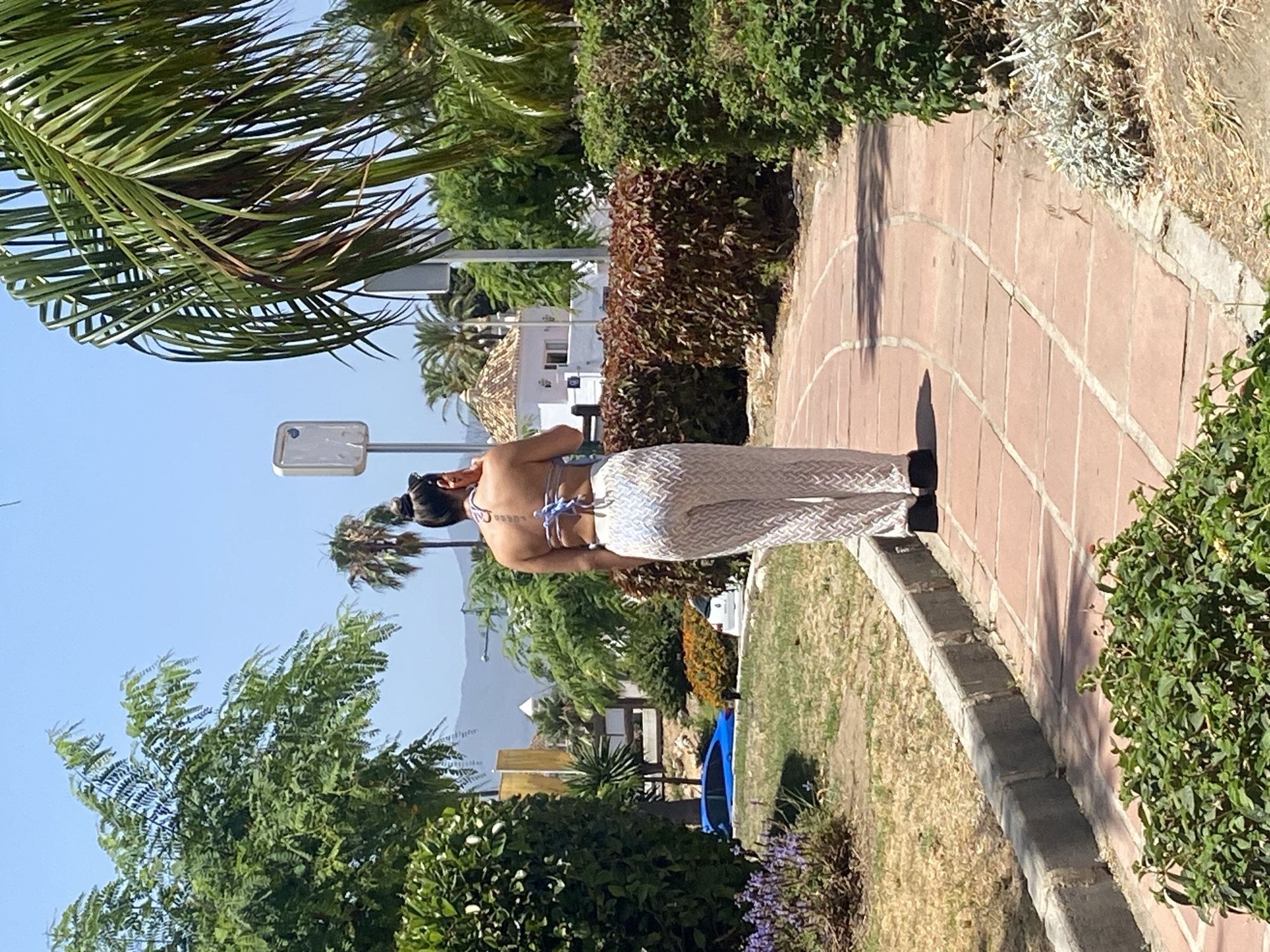 A woman with her hair in a bun, wearing a bikini top and patterned wide-leg pants, stands on a curved brick pathway. She is facing away from the camera and appears to be on her phone. The pathway winds through a green, well-maintained park with lush foliage, bushes, and some dry grass. In the background, palm trees, other greenery, and white buildings with tiled roofs are visible. A blue car is parked to the left of the pathway. The scene is illuminated by bright sunlight, suggesting a warm, possibly summer day. The location is identified as Estepona, Spain, an area known for its coastal scenery and Mediterranean climate.