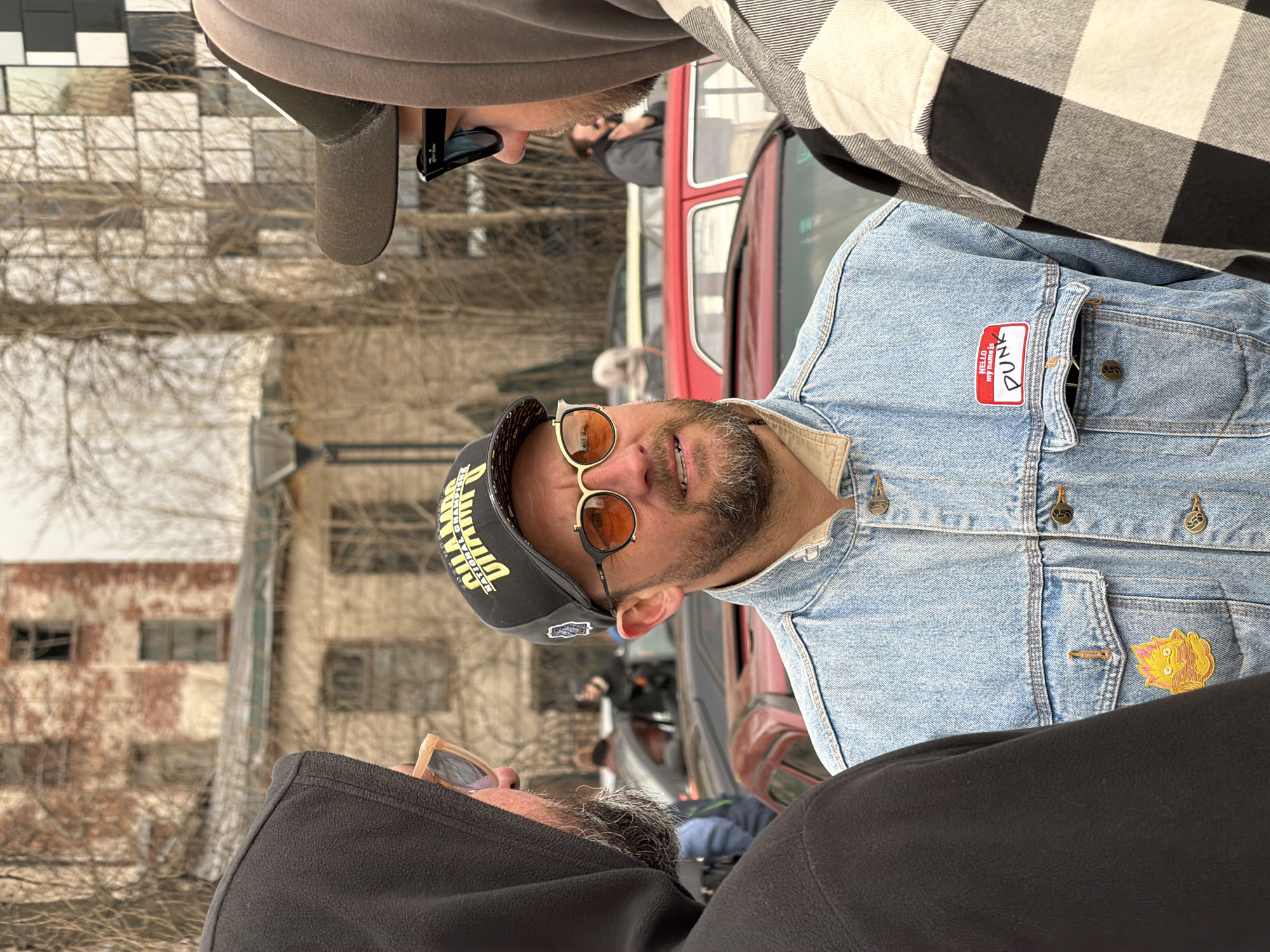 The image appears to be taken outdoors, likely on a street. In the foreground, there are three people, with the central figure wearing a denim jacket, a black hat with yellow lettering, and orange-tinted glasses. His denim jacket has a "Hello My Name Is Punk" nametag. To his right is a person wearing a black and white checkered shirt. To his left, there's a person wearing a dark hoodie and clear glasses.
Behind the foreground figures, there is a red double-decker bus. In the background, there is a building with a worn exterior and what appears to be a barren tree.
Given the presence of a double-decker bus and a potentially older building, it is not possible to confirm if the location is in Saint Petersburg, Russia, as the image doesn't provide specific landmarks or architectural elements unique to that city.