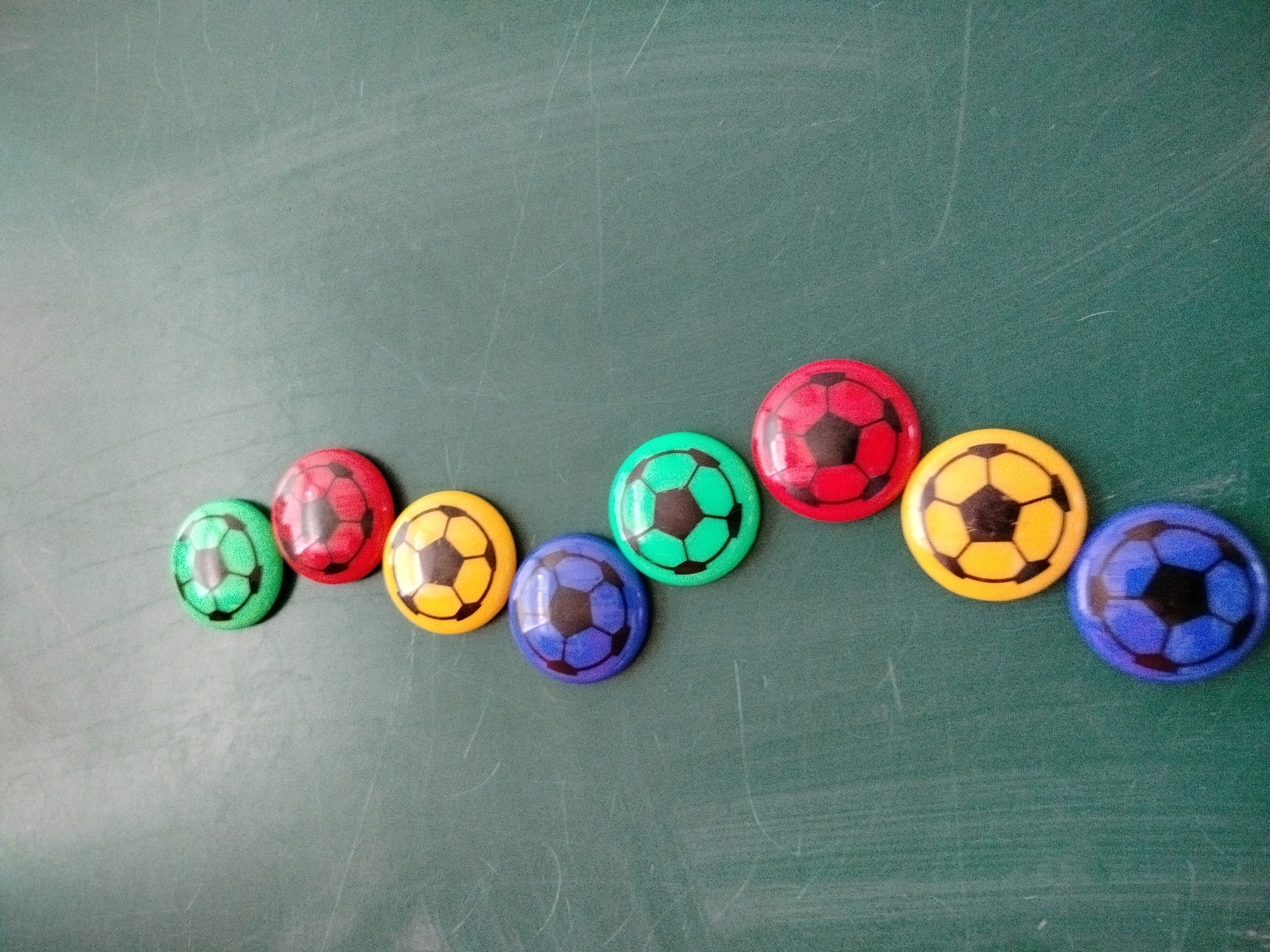 The image shows a line of nine colorful, miniature soccer balls arranged diagonally on a dark green, textured surface that appears to be a chalkboard. The balls are not in a perfectly straight line, but rather form a slightly wavy pattern from the bottom left to the top right. From left to right, the colors of the balls are: green, red, yellow, green, blue, red, yellow, blue, and blue. Each ball has a black pentagonal pattern printed on it, mimicking the design of a traditional soccer ball. The lighting suggests the image was taken in a well-lit indoor environment, with some slight glare visible on the surfaces of the balls. The background is a uniform dark green, with faint horizontal and vertical scratch marks visible, characteristic of a chalkboard. There are no people or other objects visible in the frame. The location in the city of Көлкент, Kazakhstan is not determinable from the visual information present in the image.