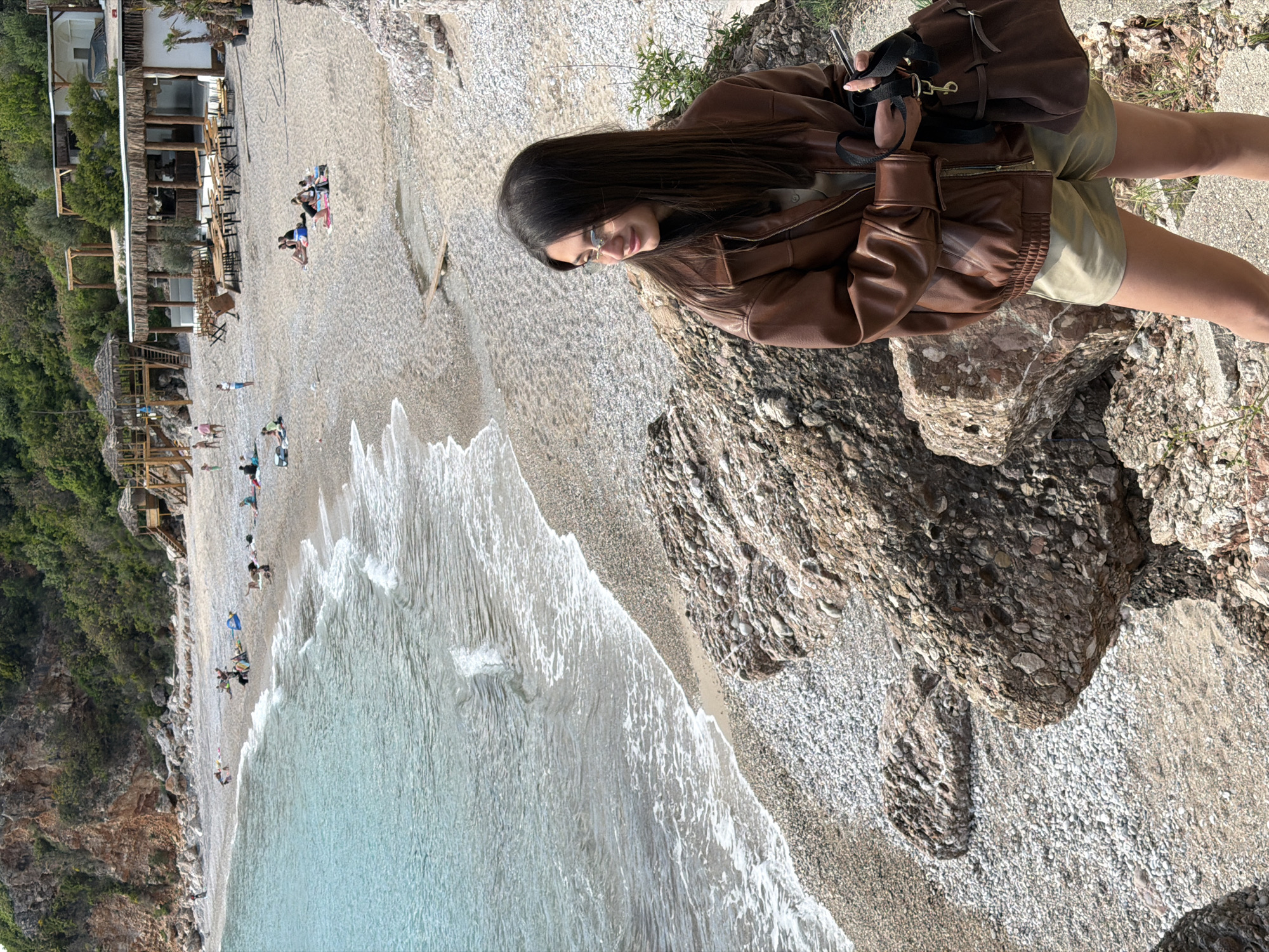 A woman stands on a rocky cliff overlooking a beach in Drobnići, Montenegro. The woman is wearing a brown leather jacket, khaki shorts, and eyeglasses. She also has a brown backpack. Below her is the beach with several people on it, and the ocean. There is a building on a cliff to the left side of the beach. The environment is sunny, and the water is a clear turquoise color.