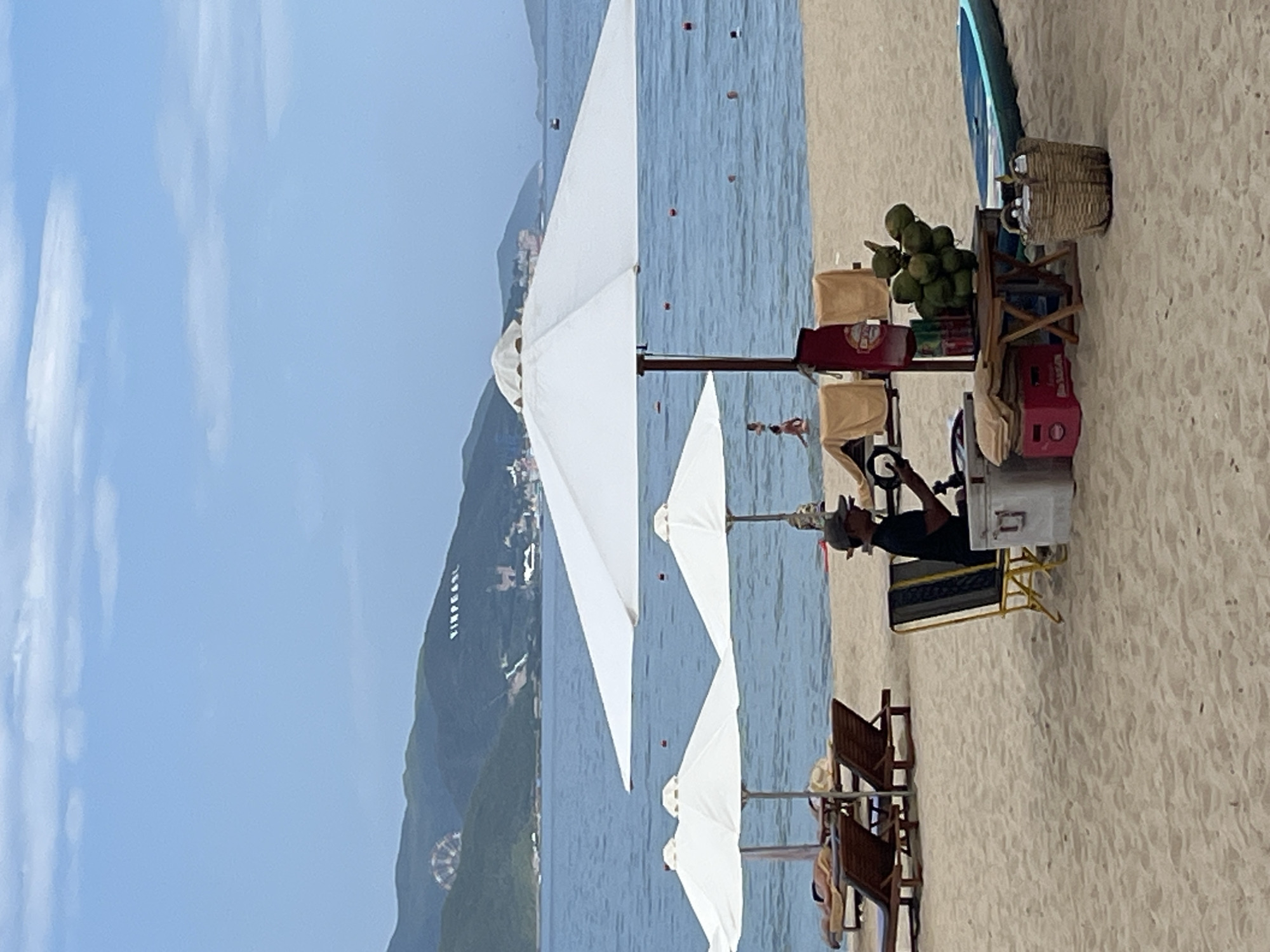 The image depicts a beach scene in Nha Trang, Vietnam, likely in the vicinity of Vinpearl Land, as indicated by white lettering on the distant hills. In the foreground, a sandy beach is occupied by beach chairs and umbrellas. A vendor is seated behind a small cart displaying coconuts and various items, with a cooler to their left. Further back, the sea is visible with several people swimming. In the background, lush green hills rise, topped with a large Ferris wheel and the Vinpearl lettering, suggesting a resort or amusement park area. The sky is a clear blue with scattered clouds.