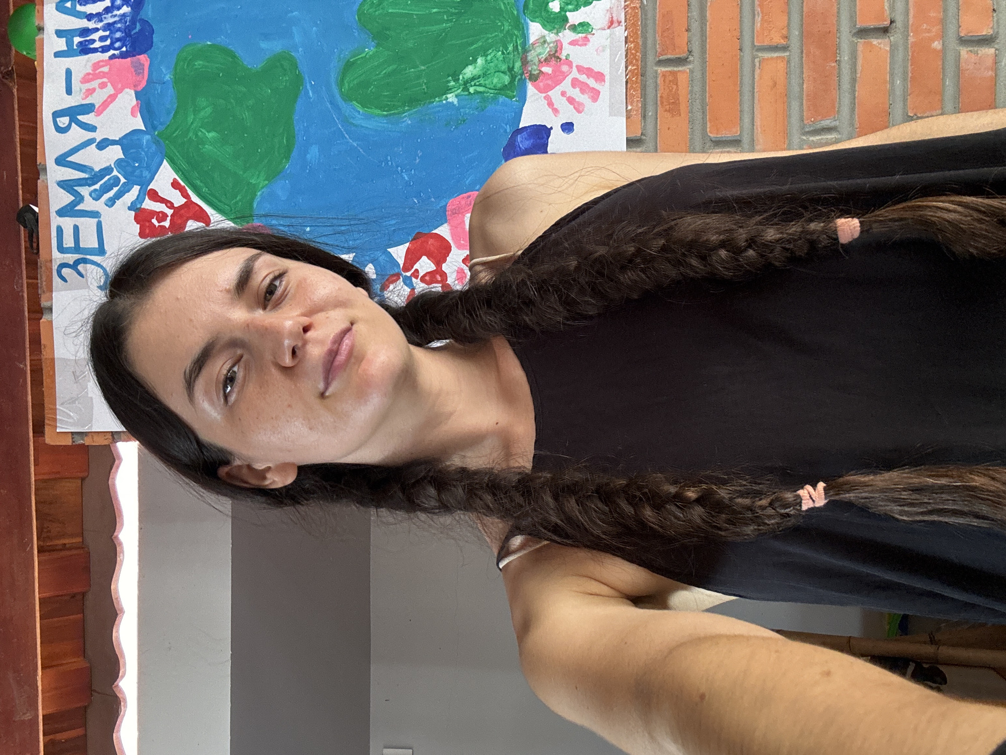 A medium-shot captures a light-skinned person with long, dark hair styled in two braids, each secured with a light pink hair tie. The person is wearing a black tank top. A painted banner hangs in the background. The banner has the word "ЗЕМЛЯ" and the phrase "Я ЛЮБЛЮ ТЕБЯ" which is Cyrillic for "I love you" painted on it, along with an image of Earth depicted in green and blue. Red and blue handprints also adorn the banner. Behind the banner, a brick wall is visible. The location is Mui Ne, Vietnam.
