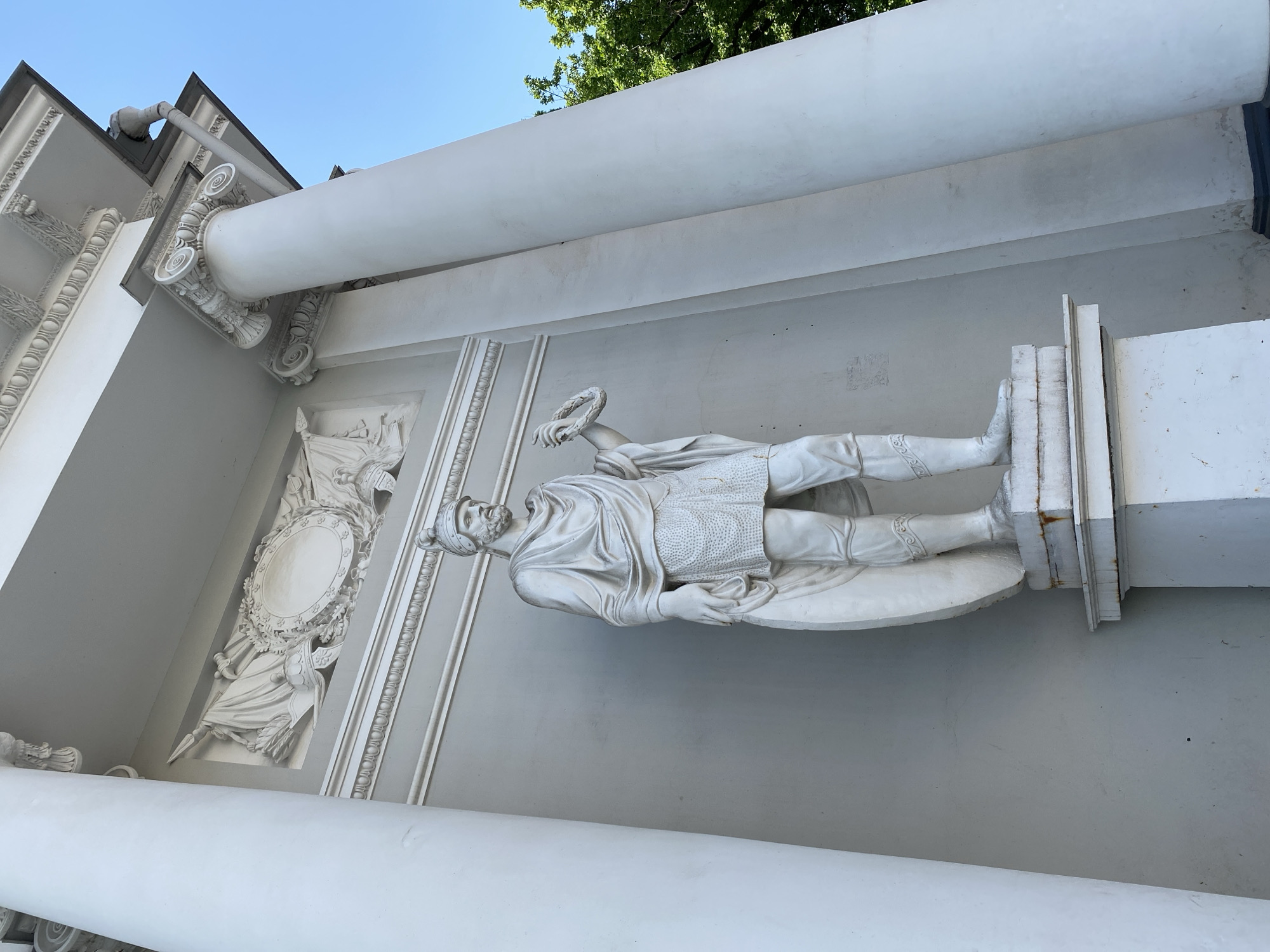 The photograph captures a white stone statue of a Roman soldier standing between two large white columns, part of a neoclassical architectural structure. The statue depicts a male figure with a beard, wearing a helmet, a draped garment over a patterned tunic, and ornate greaves on his legs. He holds a circular shield in his left hand and a wreath in his right. The sculpture is placed on a tiered pedestal. Above and behind the statue, a decorative frieze with relief carvings of flags, a shield, and a wreath adorns the wall. The architecture features classical elements like Corinthian capitals on the columns. Lush green trees are visible on the right side of the image, with a clear blue sky overhead. This structure is the monument to Prince Potemkin in Tauride Garden, Saint Petersburg, Russia.