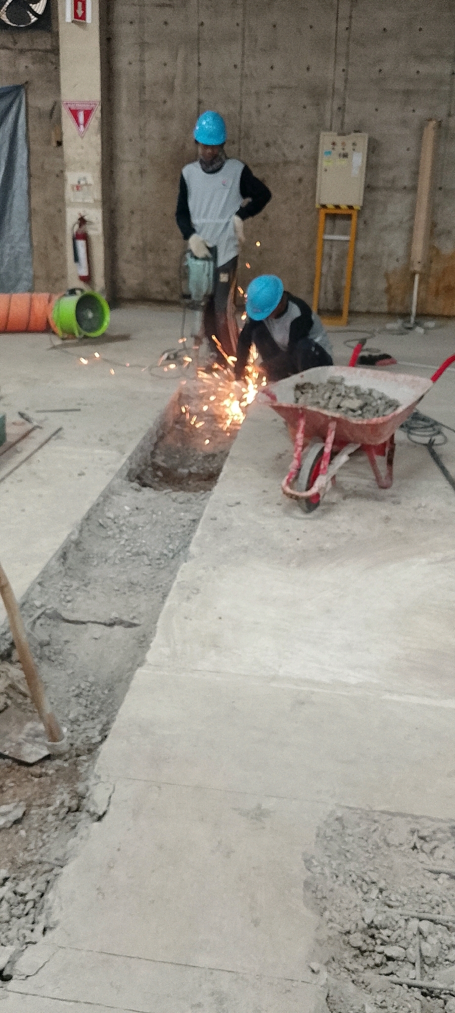 The image depicts an interior construction site, possibly within a building under development or renovation, in Depok, Indonesia. Two construction workers, both wearing blue hard hats and protective gloves, are engaged in their tasks. One worker, standing and holding a jackhammer, is in the process of breaking up concrete. Sparks are flying from the area where the jackhammer is being used, indicating active demolition or cutting. Another worker is crouched down, also working on the concrete, and is the source of a significant shower of sparks, suggesting they are using a grinder or a similar cutting tool.
In the foreground, a red wheelbarrow is filled with construction debris, consisting of broken concrete pieces. To the left of the workers, a green industrial fan is visible, possibly used for ventilation. Further back, a fire extinguisher is mounted on a wall, a safety precaution common in such environments. The walls appear to be concrete, characteristic of construction sites. A yellow metal frame holding an electrical control panel is also visible against the wall in the background. The floor is a mixture of concrete and exposed aggregate, with a trench dug into it where the work is concentrated. A wooden tool handle is seen sticking out of the ground near the trench. The overall scene conveys an active and industrious work environment.