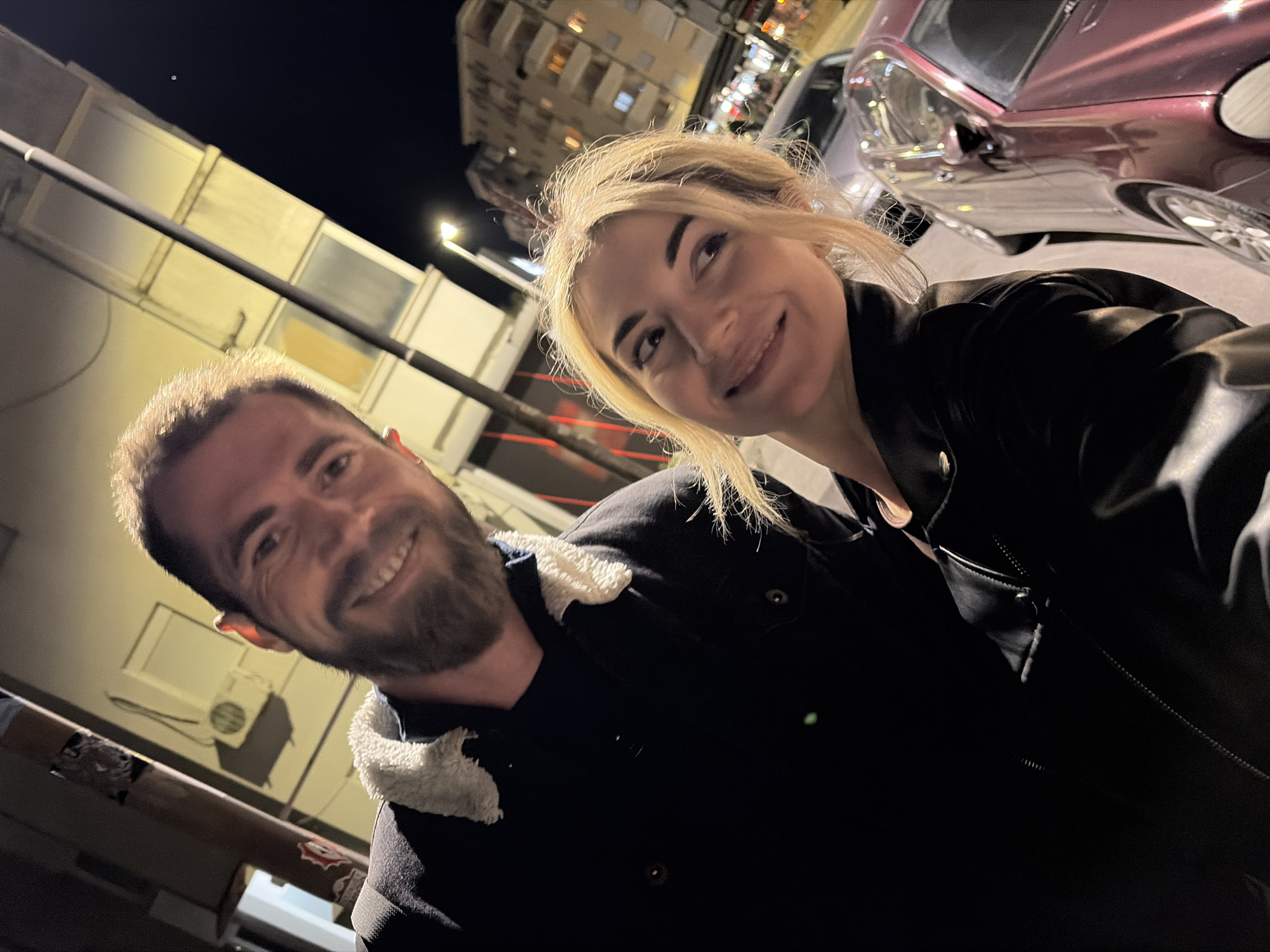 The image depicts an adult male and an adult female positioned closely in the foreground of a nighttime urban street in Budva, Montenegro. The male, on the left, has short dark hair and a full beard, wearing a dark jacket with a white shearling collar, and is smiling directly towards the camera. The female, on the right, has blonde hair and wears a dark jacket with a visible zipper. She is also smiling, with her gaze directed slightly to the right of the camera. The background reveals building facades, one with an exterior air conditioning unit, a street pole, and a streetlight. Parked vehicles, including a reddish-purple sedan, are visible on the right. Multi-story buildings with illuminated windows are discernible in the distant background under a dark sky. The scene is illuminated by artificial street and building lights.